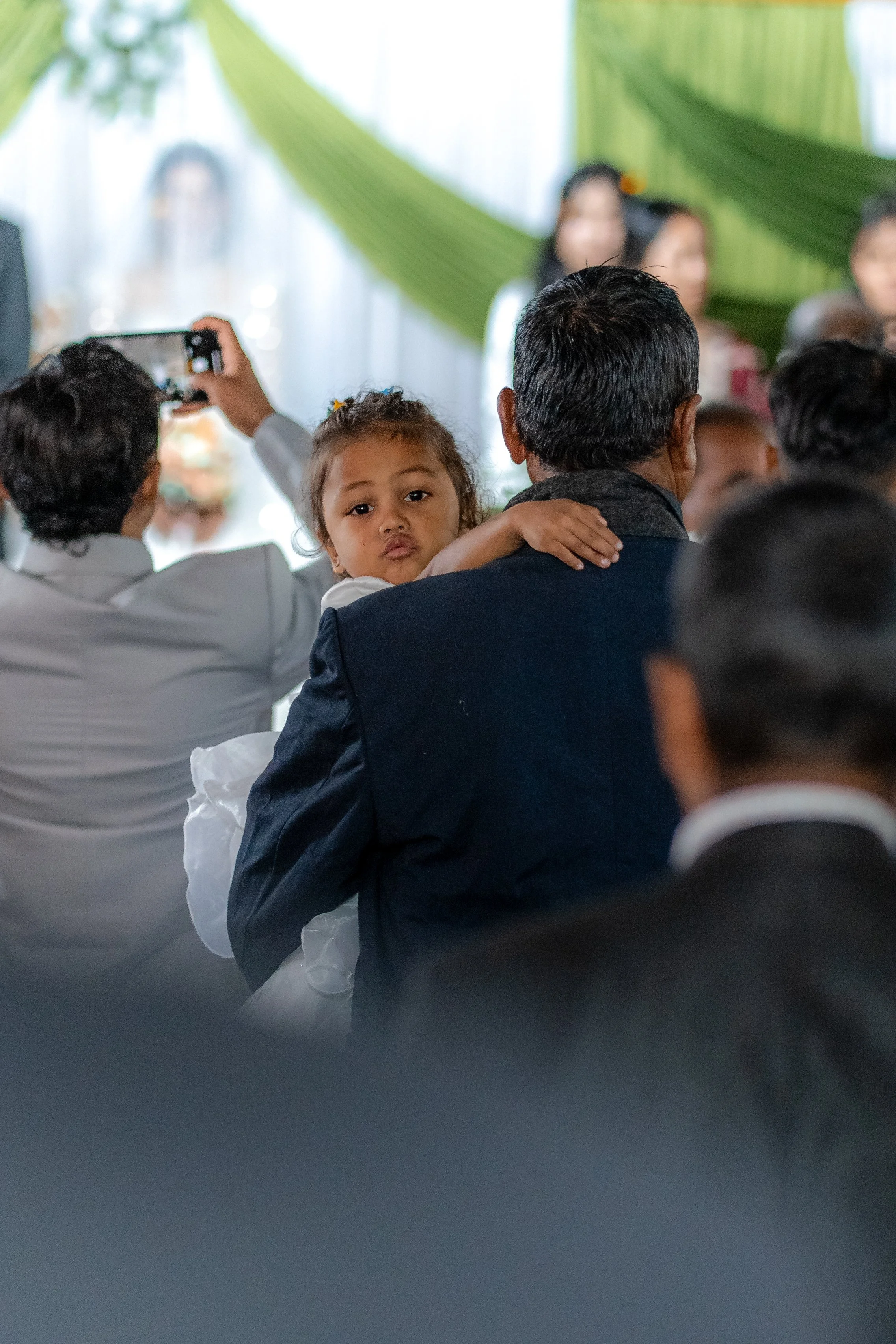A young girl with curly hair is resting her head on a man's shoulder, who is wearing a dark suit, in a crowded indoor setting with green decorations in the background. A person beside them is taking a photo on their phone.