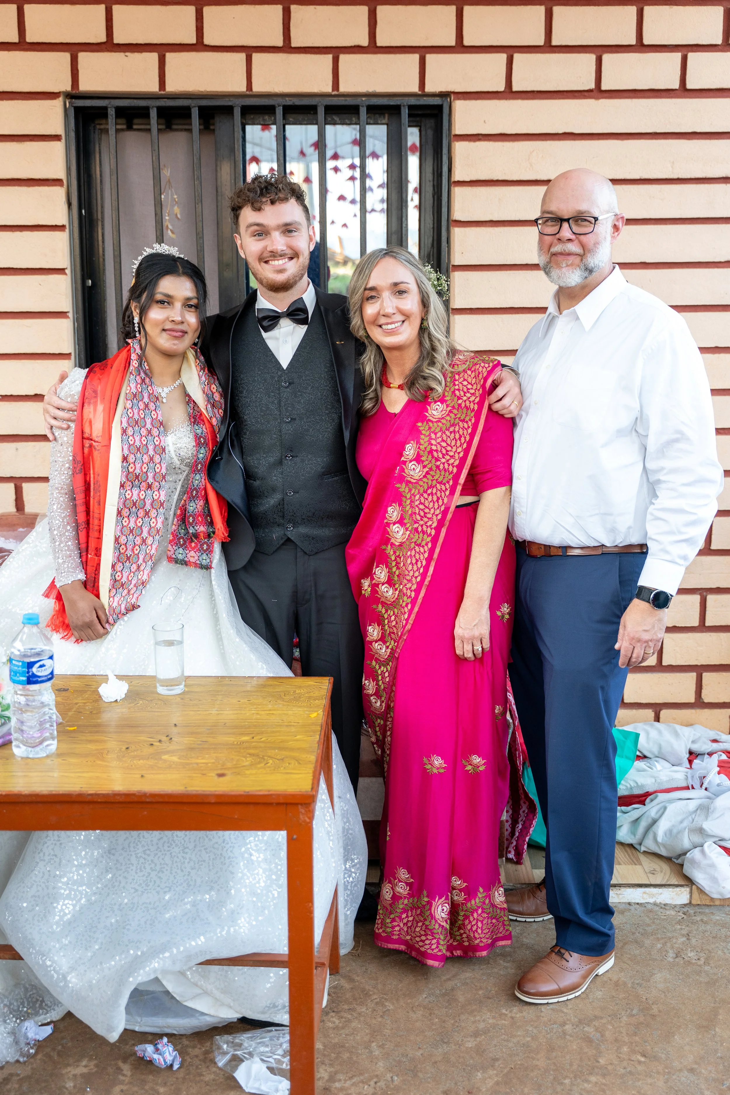 A wedding celebration with four people, including a bride in a white gown with a tiara and red scarf, a groom in a tuxedo, a woman in a vibrant pink traditional dress, and a man in a white shirt and blue pants, standing outside near a brick wall.