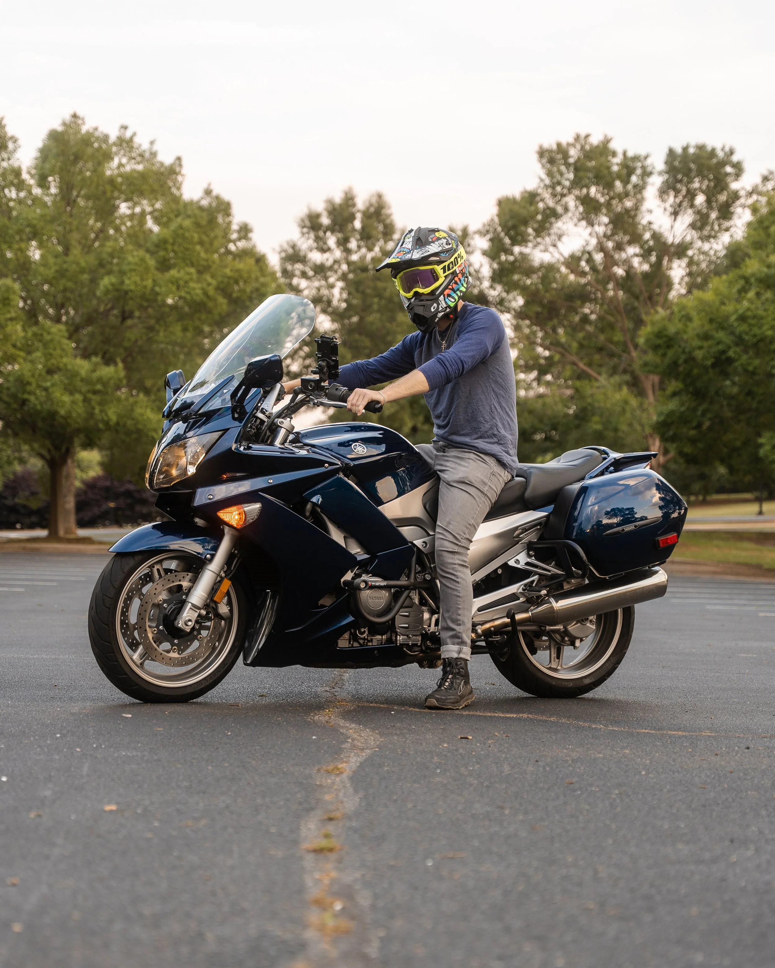 Person wearing a helmet and casual clothes sitting on a Yamaha motorcycle in an empty parking lot with trees in the background.