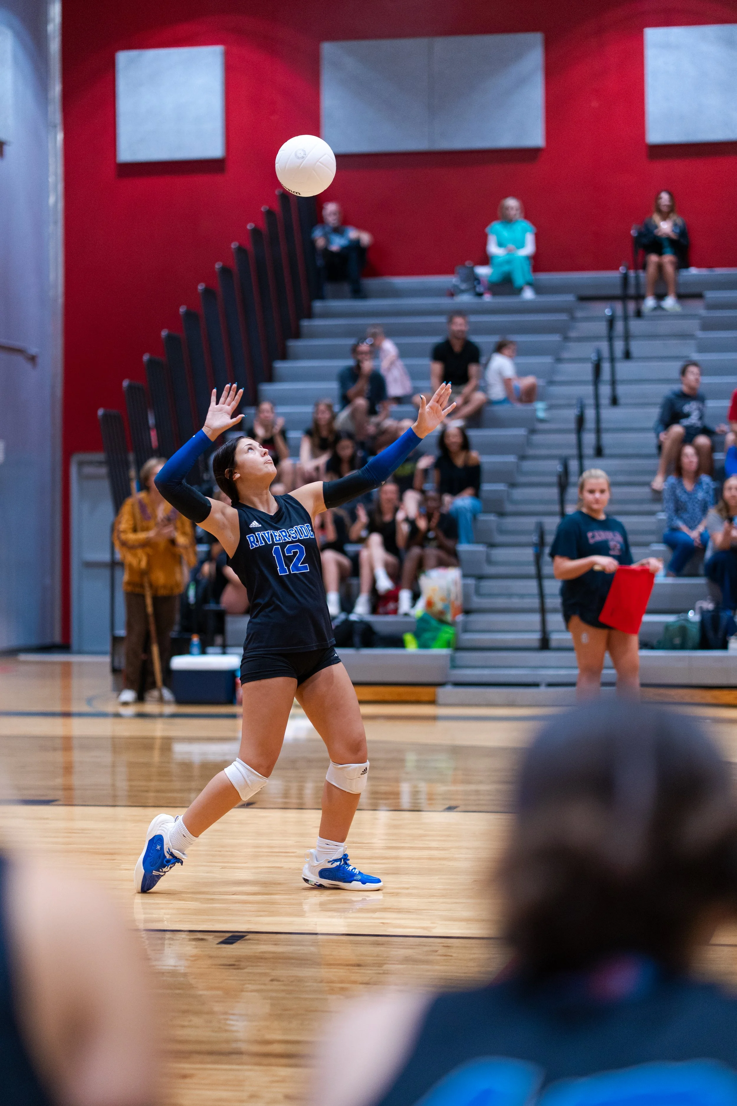 A female volleyball player wearing a black jersey with the number 12 and 'Riverside' on it, is setting a volleyball in a gymnasium with spectators watching from the bleachers.