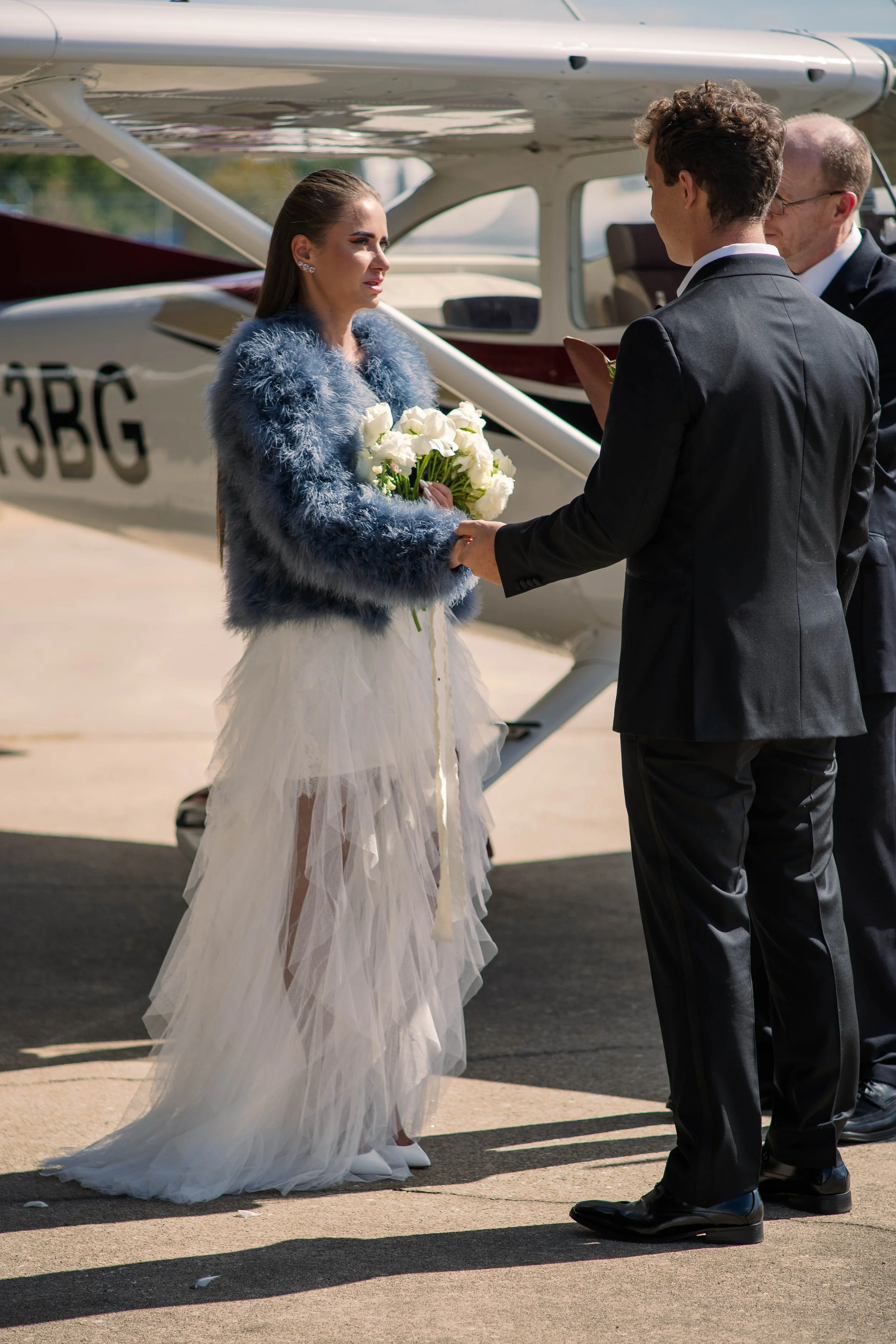 A woman in a white wedding dress with a blue fur jacket holding a bouquet of white flowers, standing in front of a small airplane, while a man in a black suit appears to be giving her a ring or a gift.