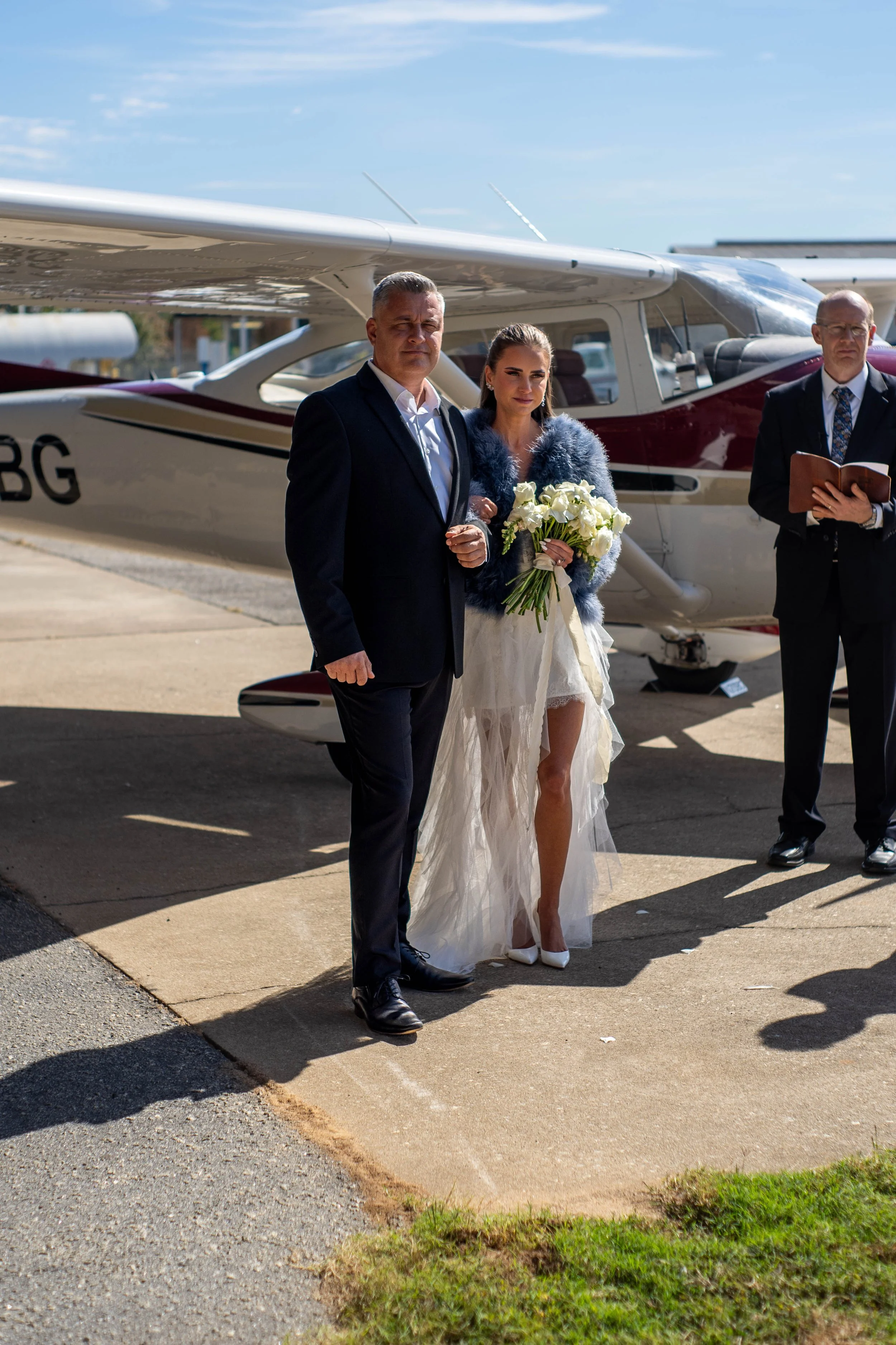 A bride and an older man, possibly her father, standing together outside near a small airplane during a wedding ceremony. The bride is holding a bouquet of white roses and wearing a white dress with a sheer train, a blue fur jacket, and white heels. 