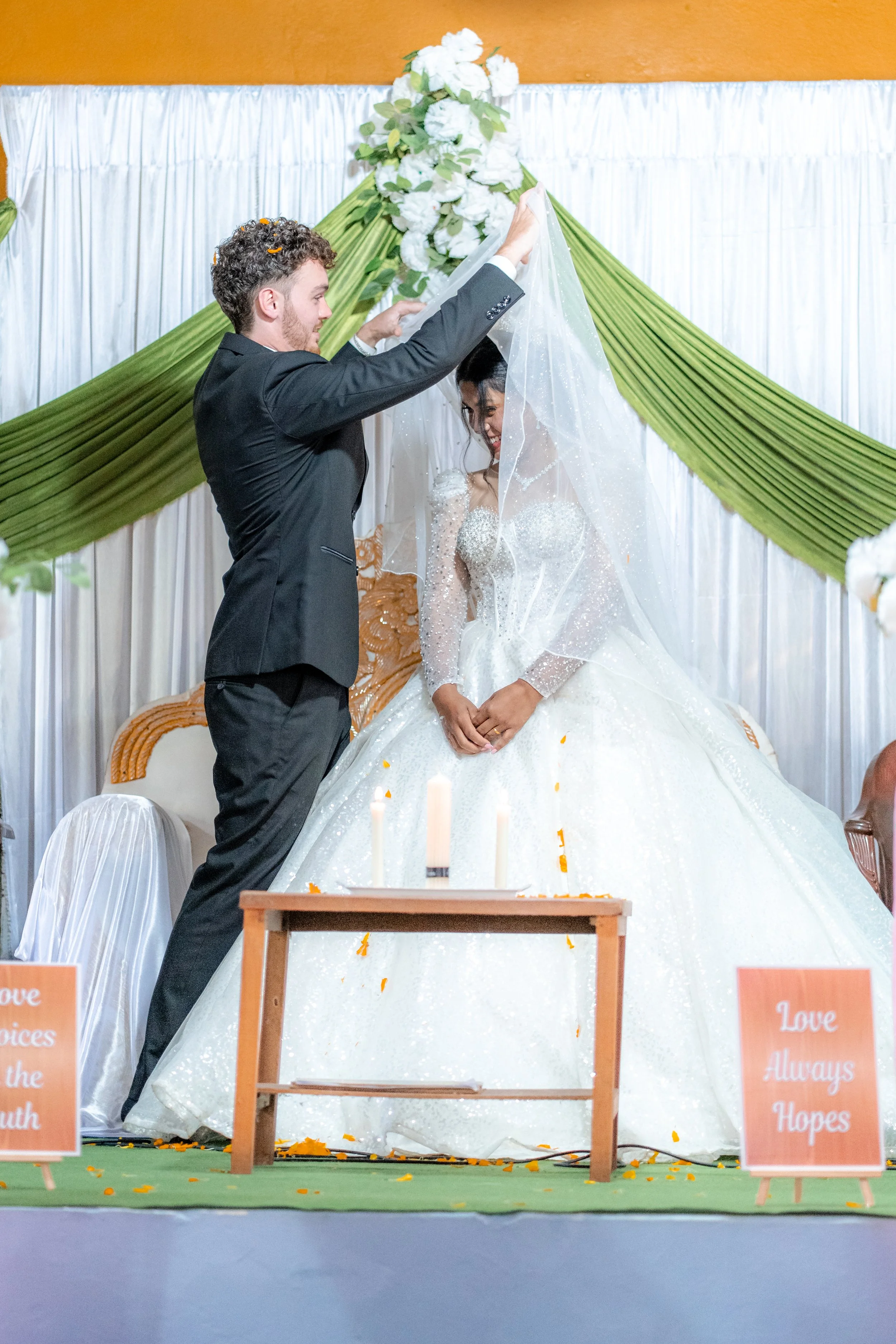 A bride and groom during their wedding ceremony. The groom is removing the veil from the bride's face. They are standing in front of a decorated backdrop with green drapes and white flowers, with candles and signs that read "Love Always Hopes."