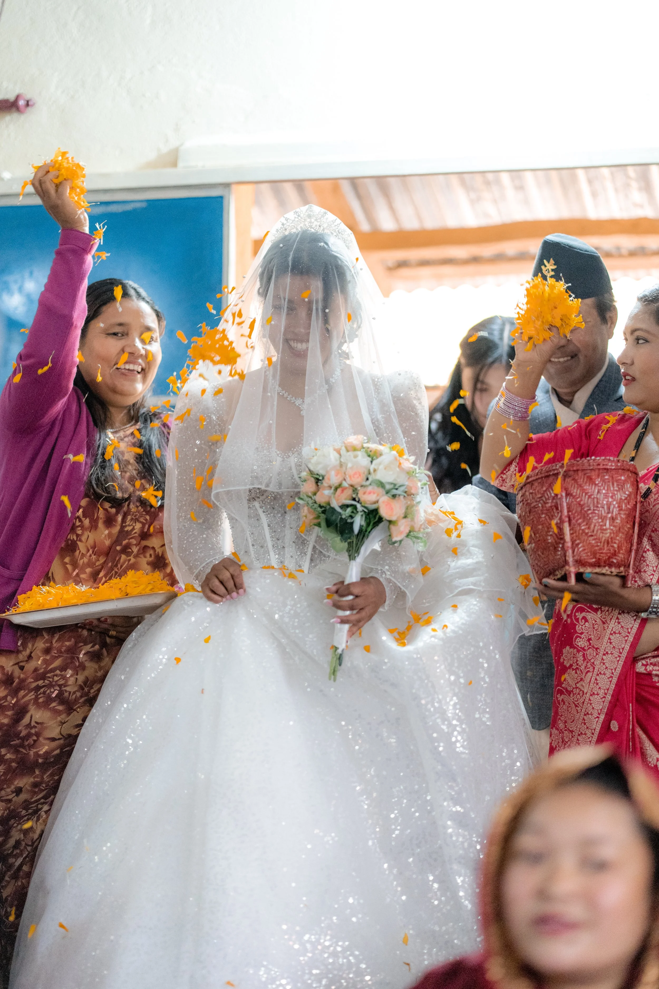 Bride in a white wedding gown holding a bouquet, surrounded by friends and family throwing orange flower petals during a celebration.