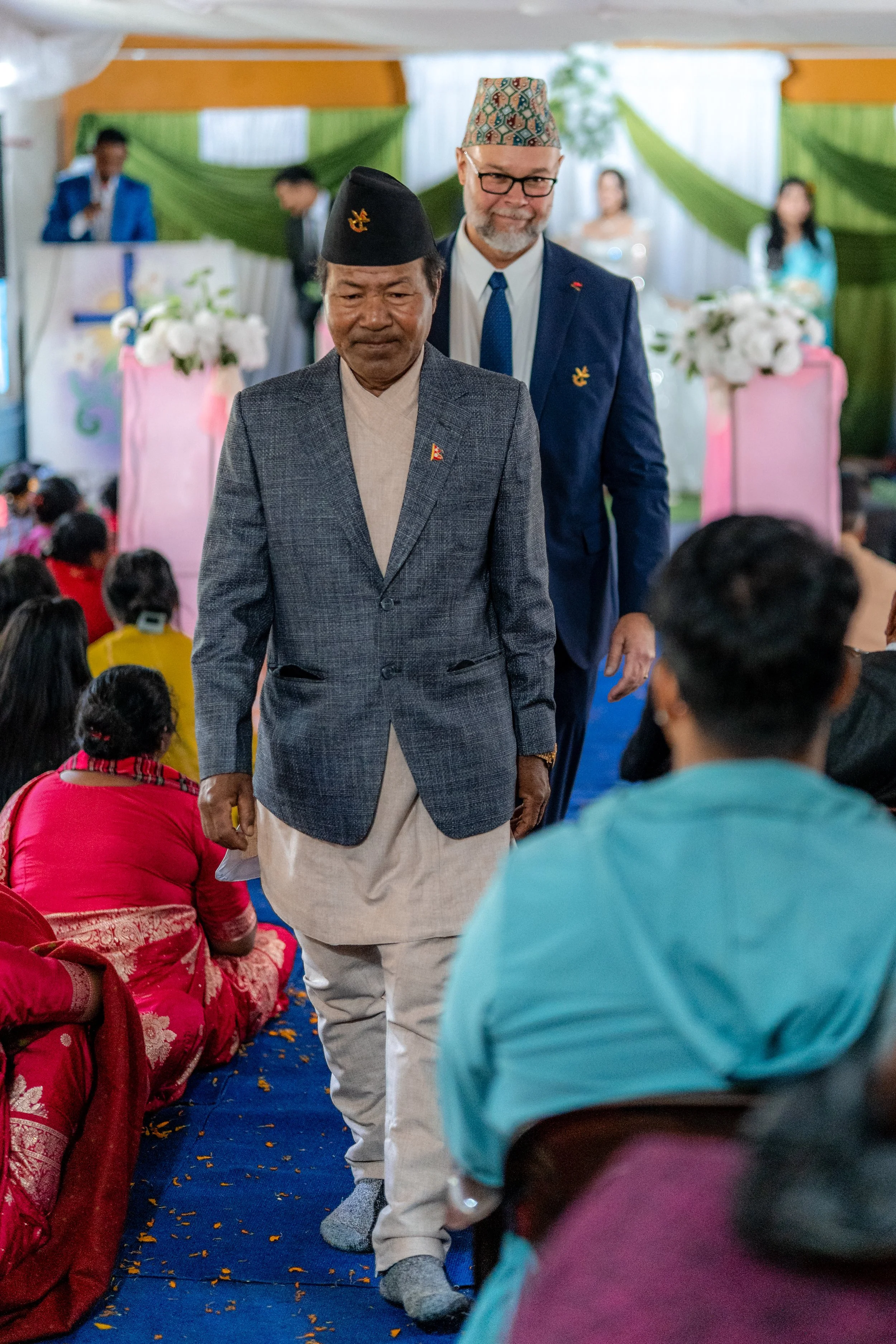 Two men, one older and one younger, walking across a seated crowd at a formal event in Nepal, with the older man in traditional Nepali attire and the younger man in a suit and glasses.