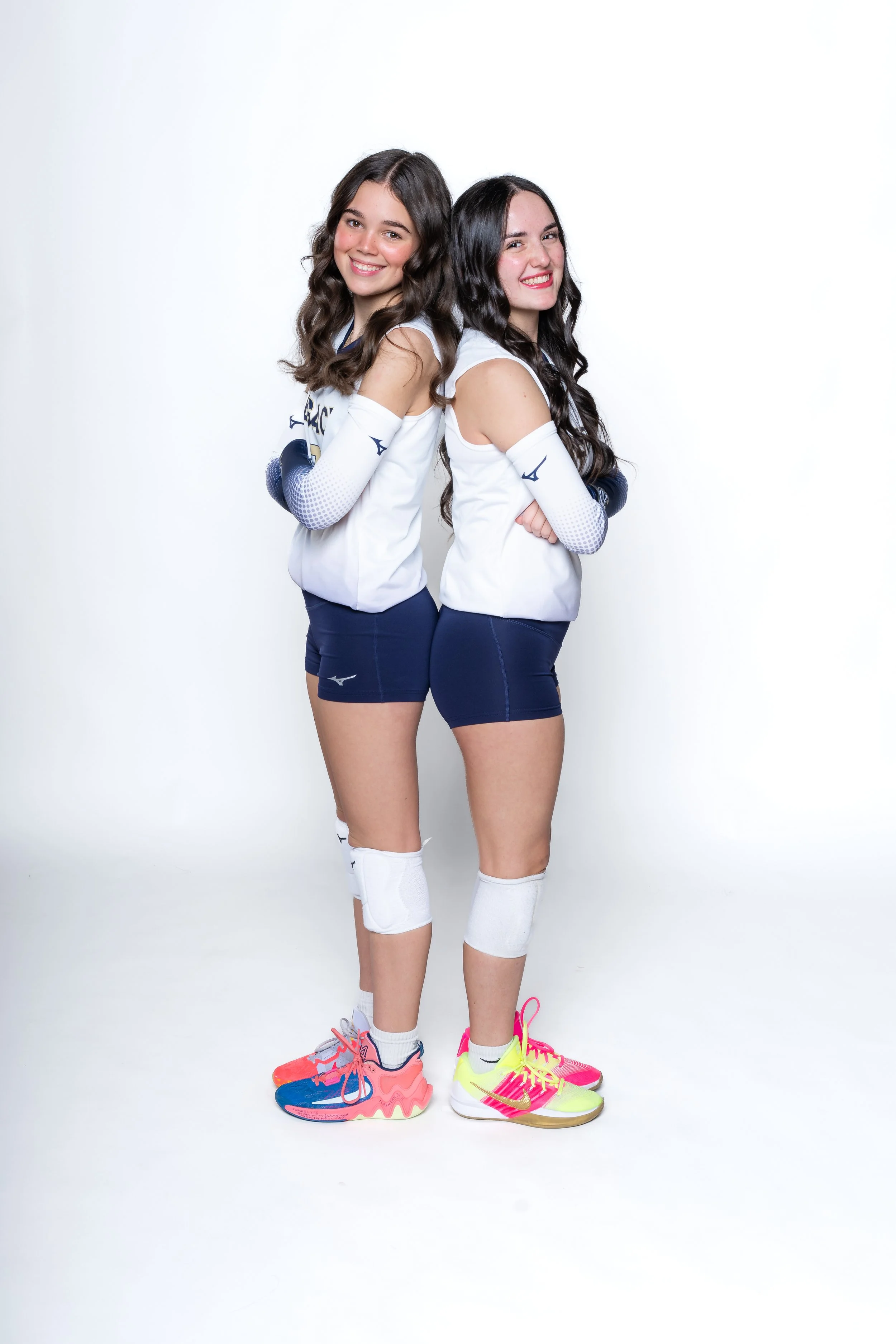 Two young women standing back-to-back wearing volleyball uniforms, with athletic shoes, knee pads, and long-sleeve arm sleeves, smiling at the camera against a white background.