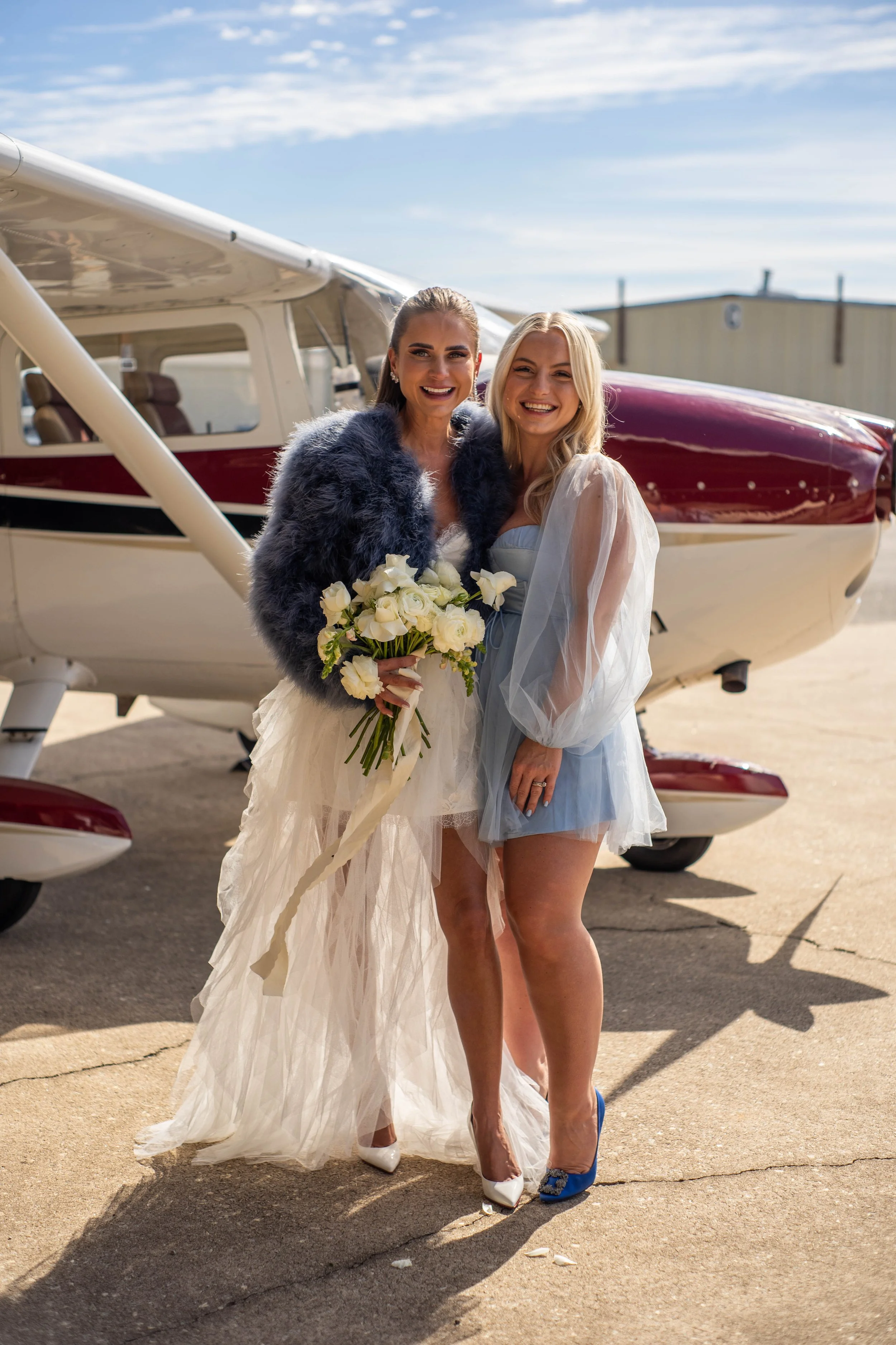 Two women in wedding attire smiling and holding a bouquet of white roses, standing in front of a small airplane outdoors on a sunny day.