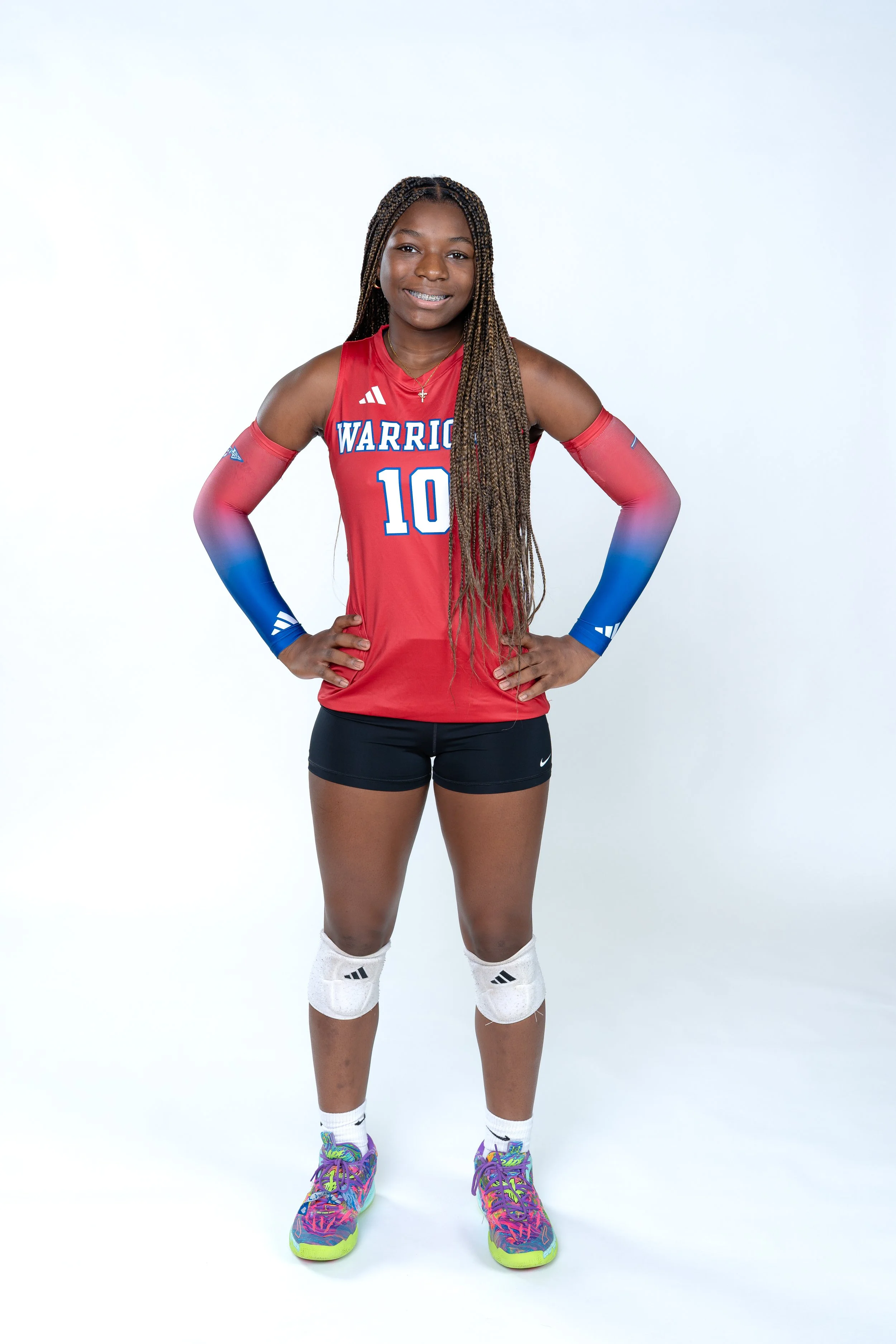 Young female volleyball player in a red and blue uniform with the number 10 and "WARRIOR" written on the front, standing with hands on hips and smiling, on a white background.