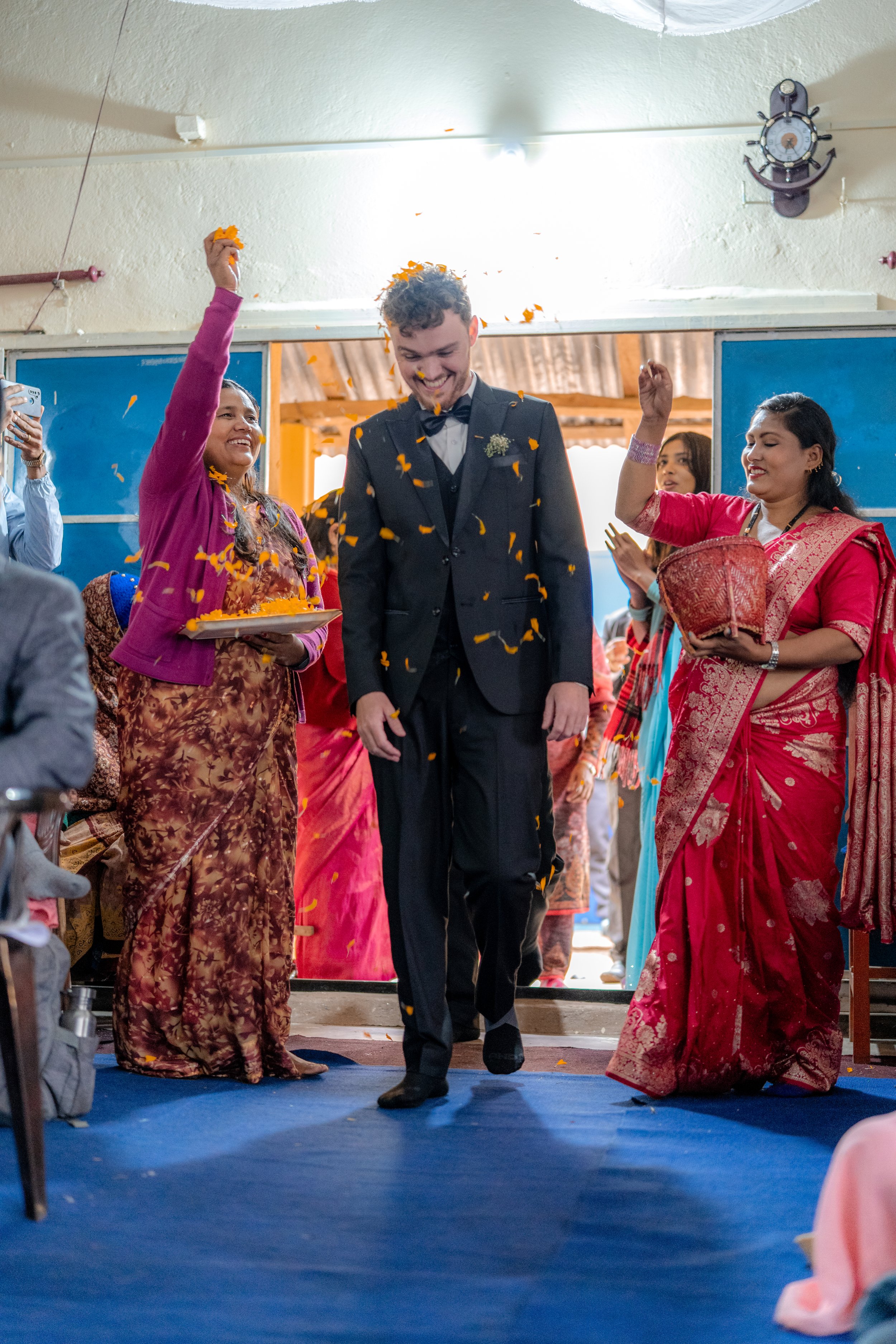 A wedding celebration with a groom walking into a room surrounded by women throwing orange flower petals.