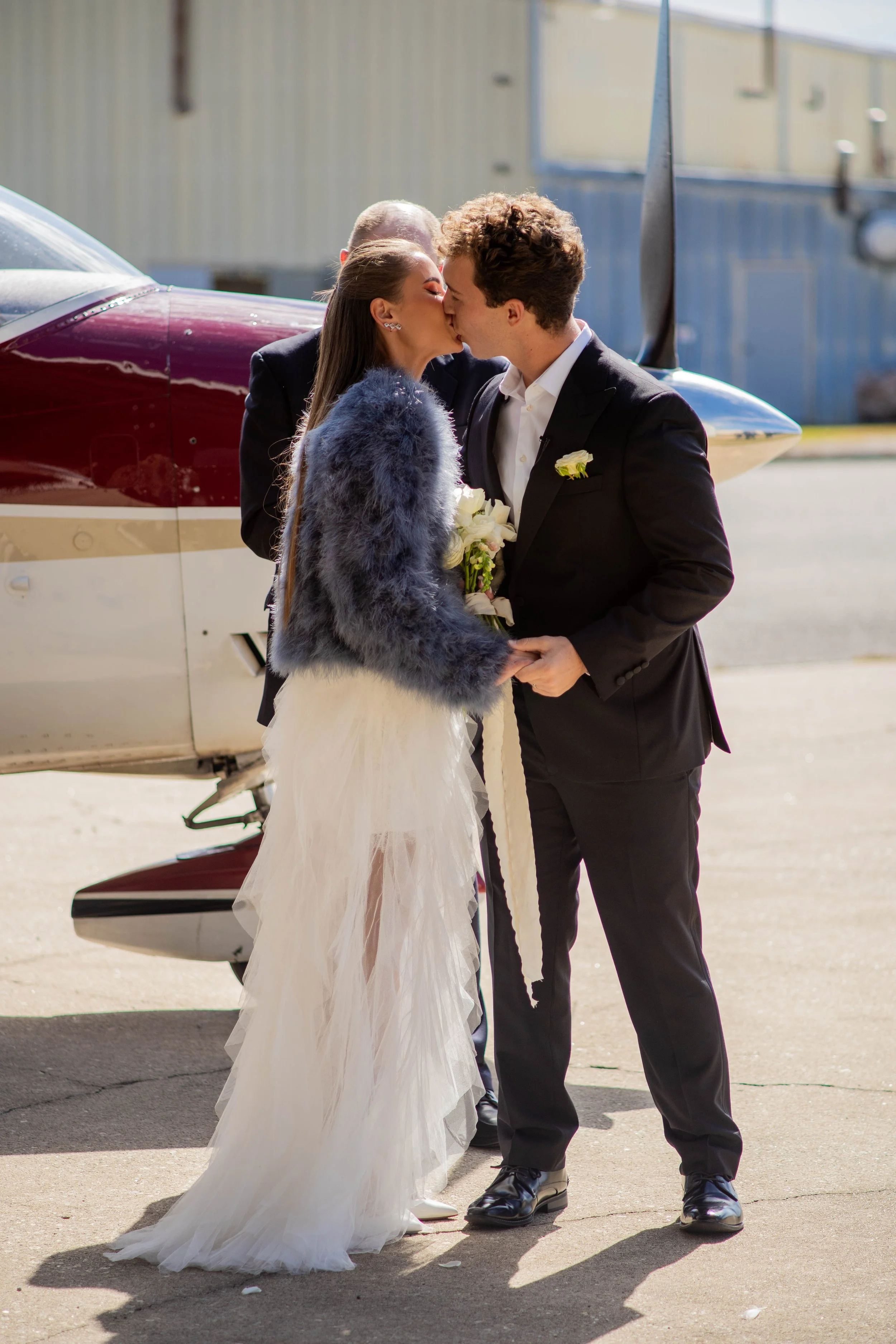 A couple in wedding attire sharing a kiss on a tarmac next to a small airplane, with a woman holding a bouquet of white flowers.