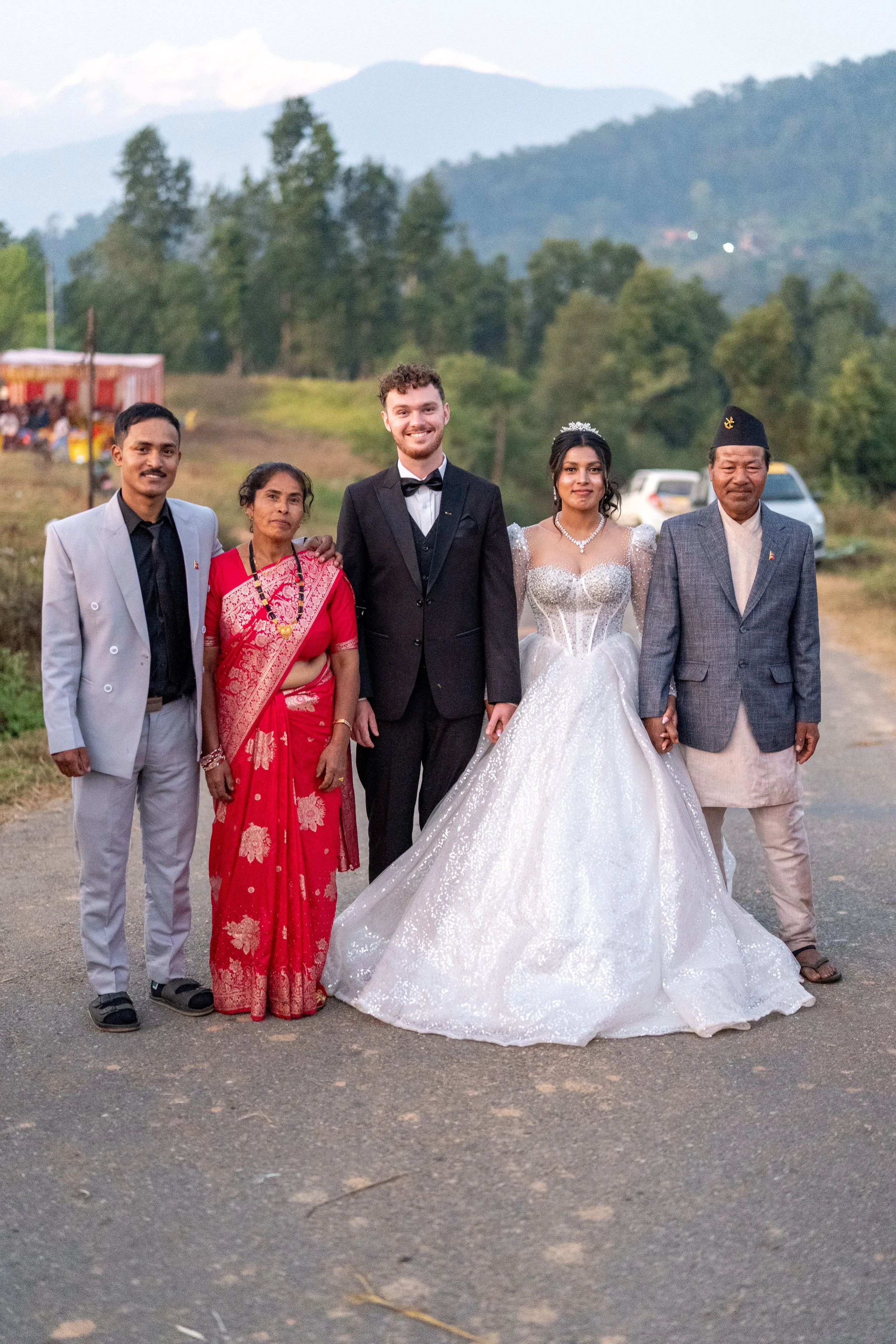 A wedding group photo with five people, including a bride in a white gown and groom in a black suit, standing outdoors on a road with green trees and mountains in the background.
