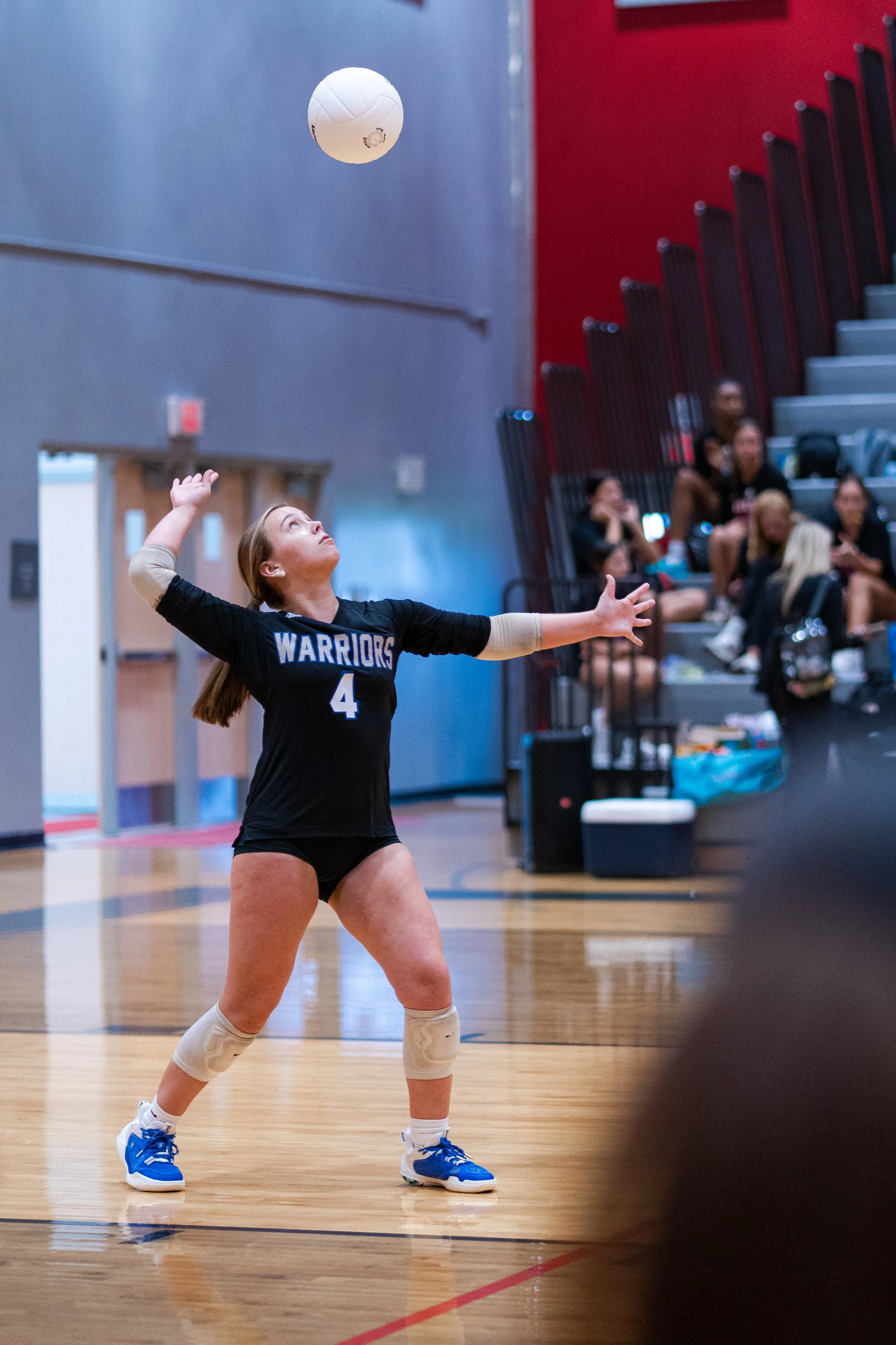 A female volleyball player in a black jersey with number 4 is setting the ball in an indoor gym. She is wearing beige knee pads and blue sneakers, with spectators sitting on bleachers in the background.