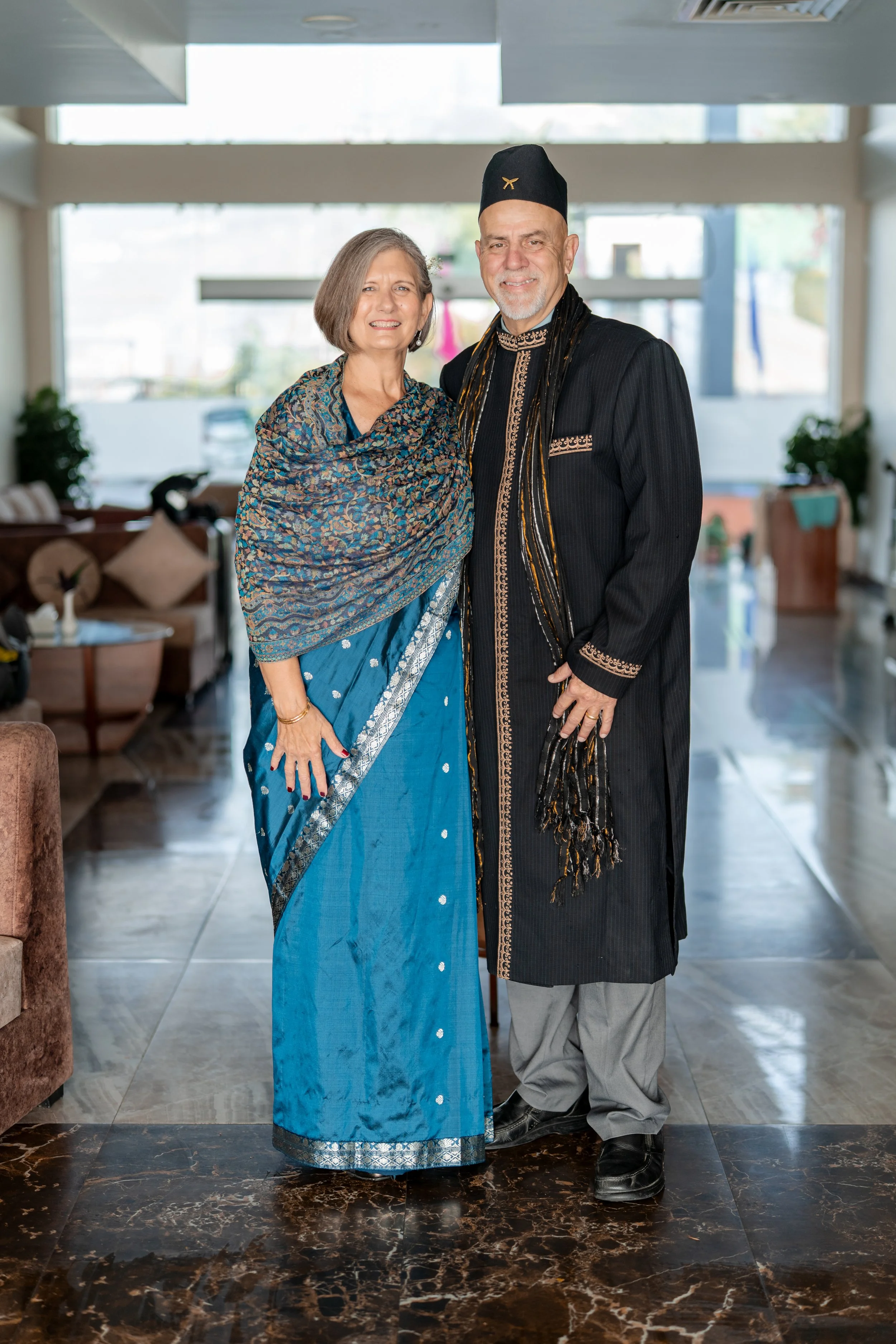 A man and woman dressed in traditional Indian attire standing indoors, smiling at the camera.