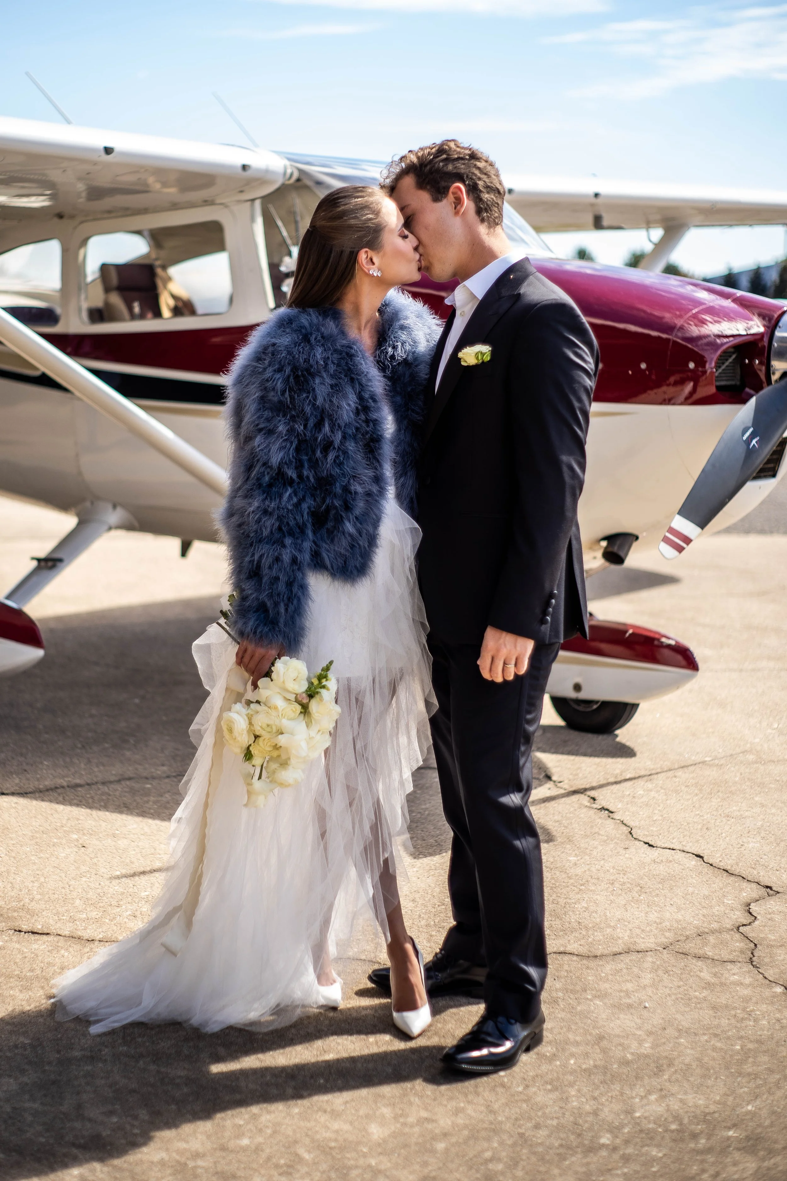 A newlywed couple kissing in front of a small airplane, the bride in a white wedding dress with a blue fur jacket and holding a bouquet of white roses, and the groom in a black suit with a white shirt, on a tarmac under a partly cloudy sky.
