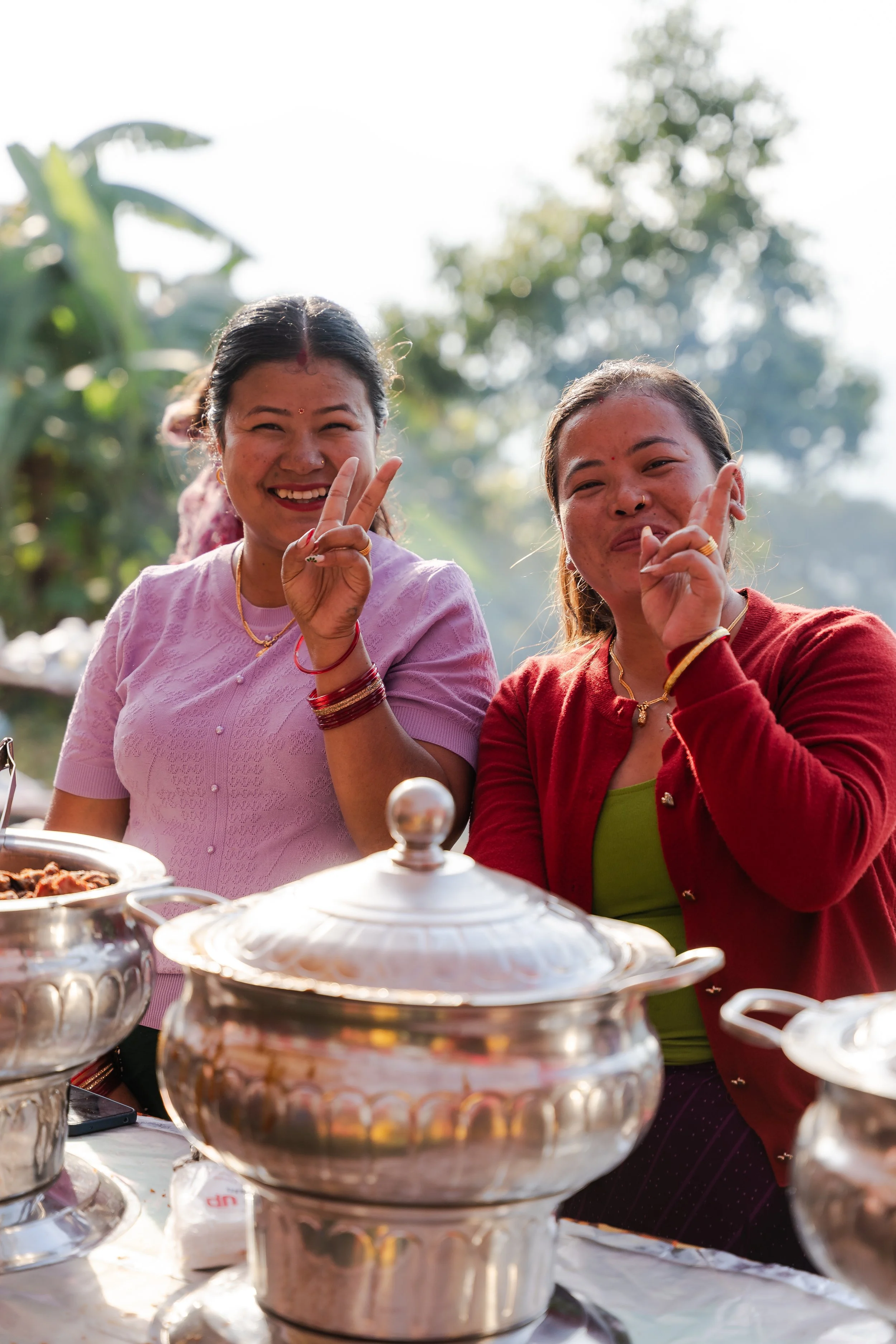 Two women smiling and making peace signs at an outdoor gathering with silver food containers on a table in front of them.