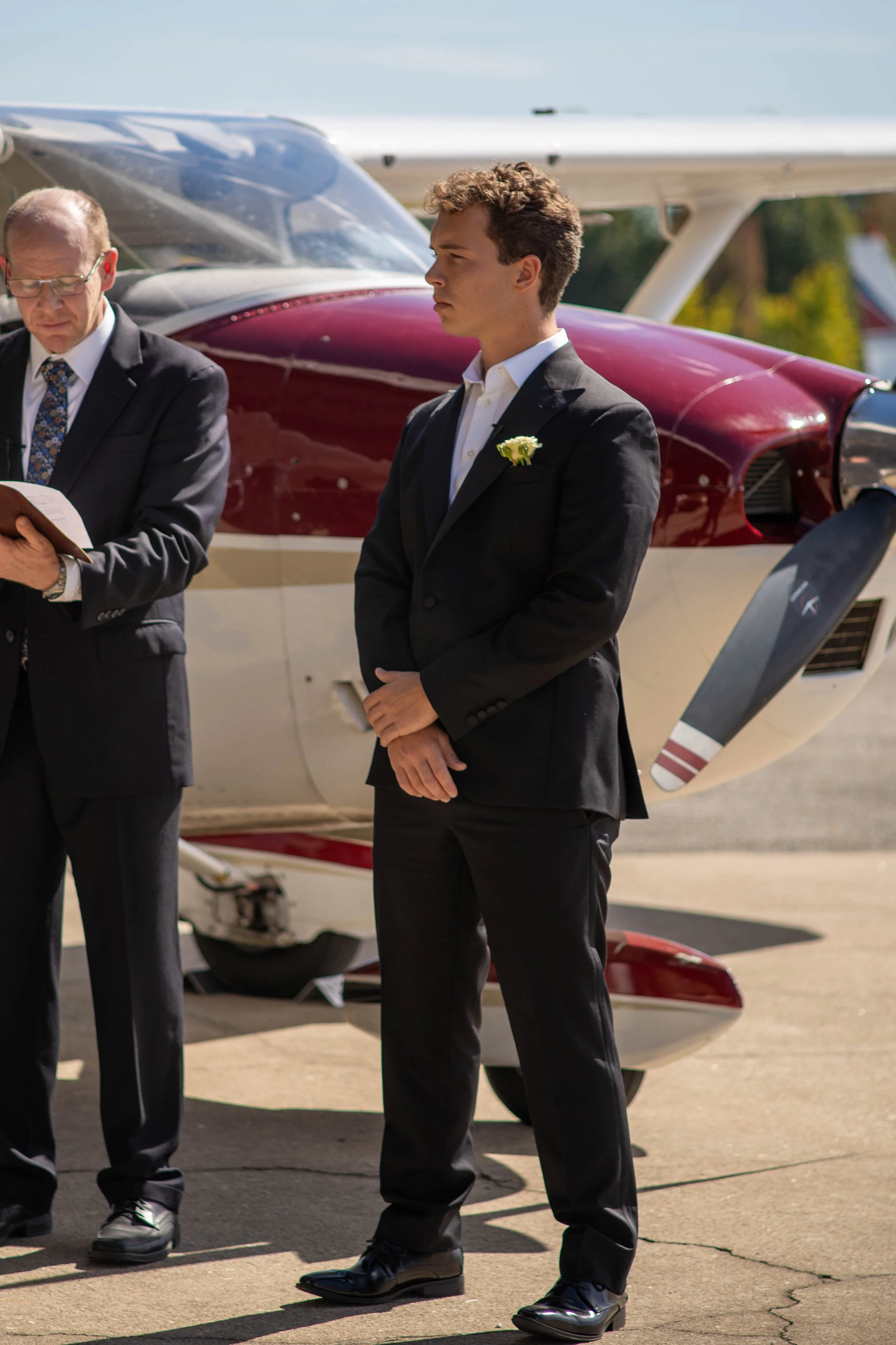 A young man in a black suit standing outdoors near a small airplane, with an older man in a suit reading from a book beside him.