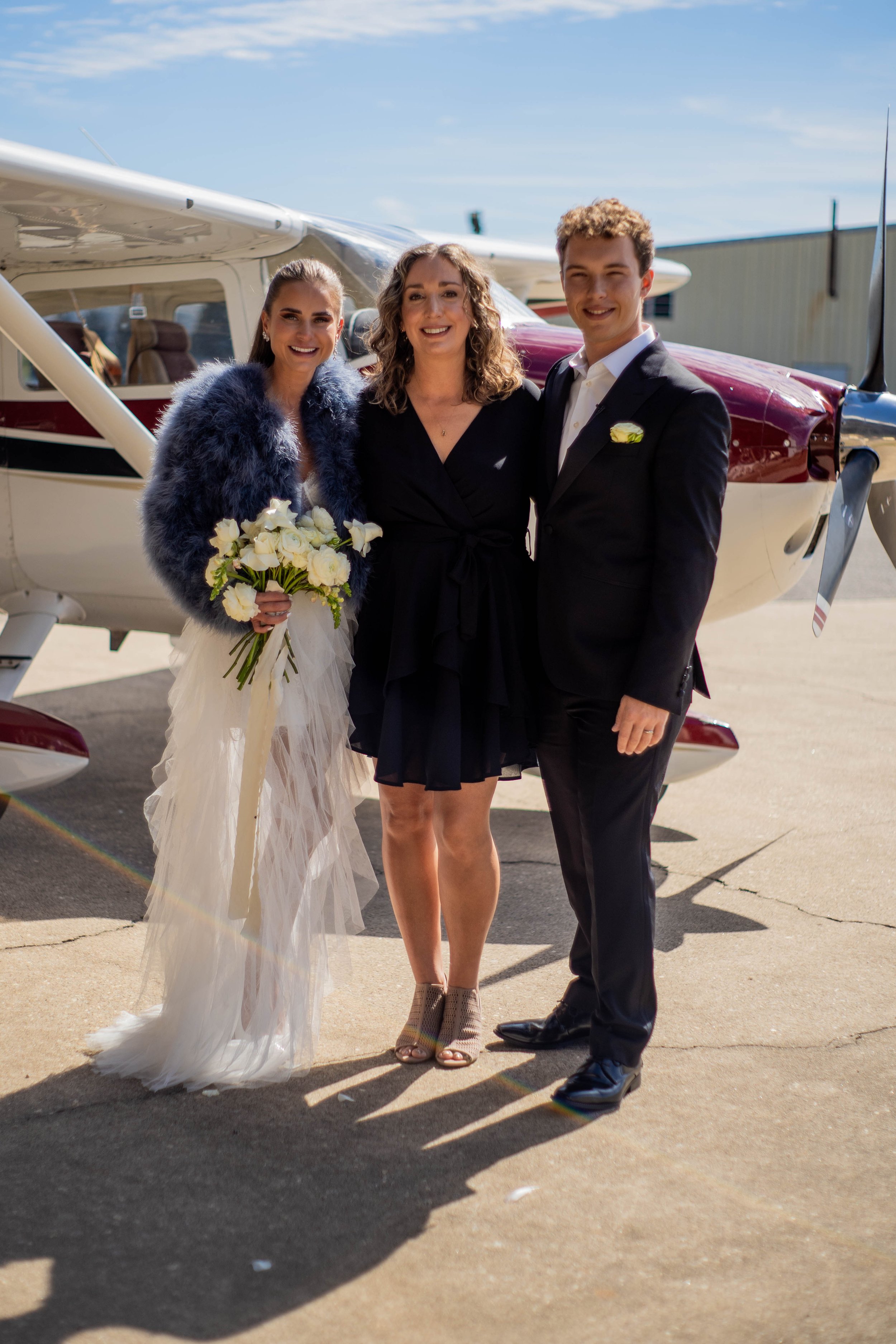 Three people standing in front of a small airplane, dressed in formal attire, smiling at the camera during daytime.