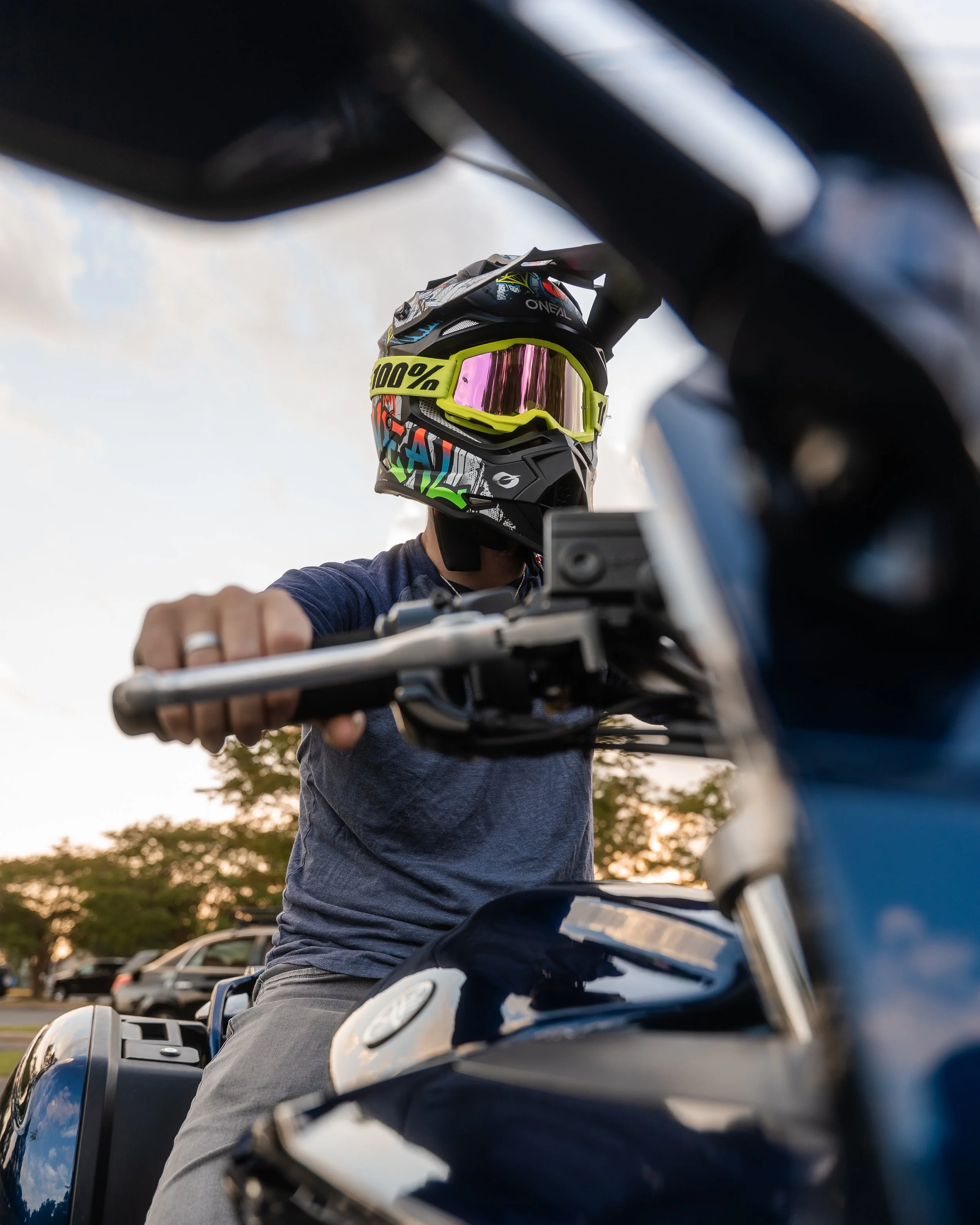 A person wearing a colorful motocross helmet with pink tinted goggles, sitting on a motorcycle, captured from below during sunset with trees and cars in the background.