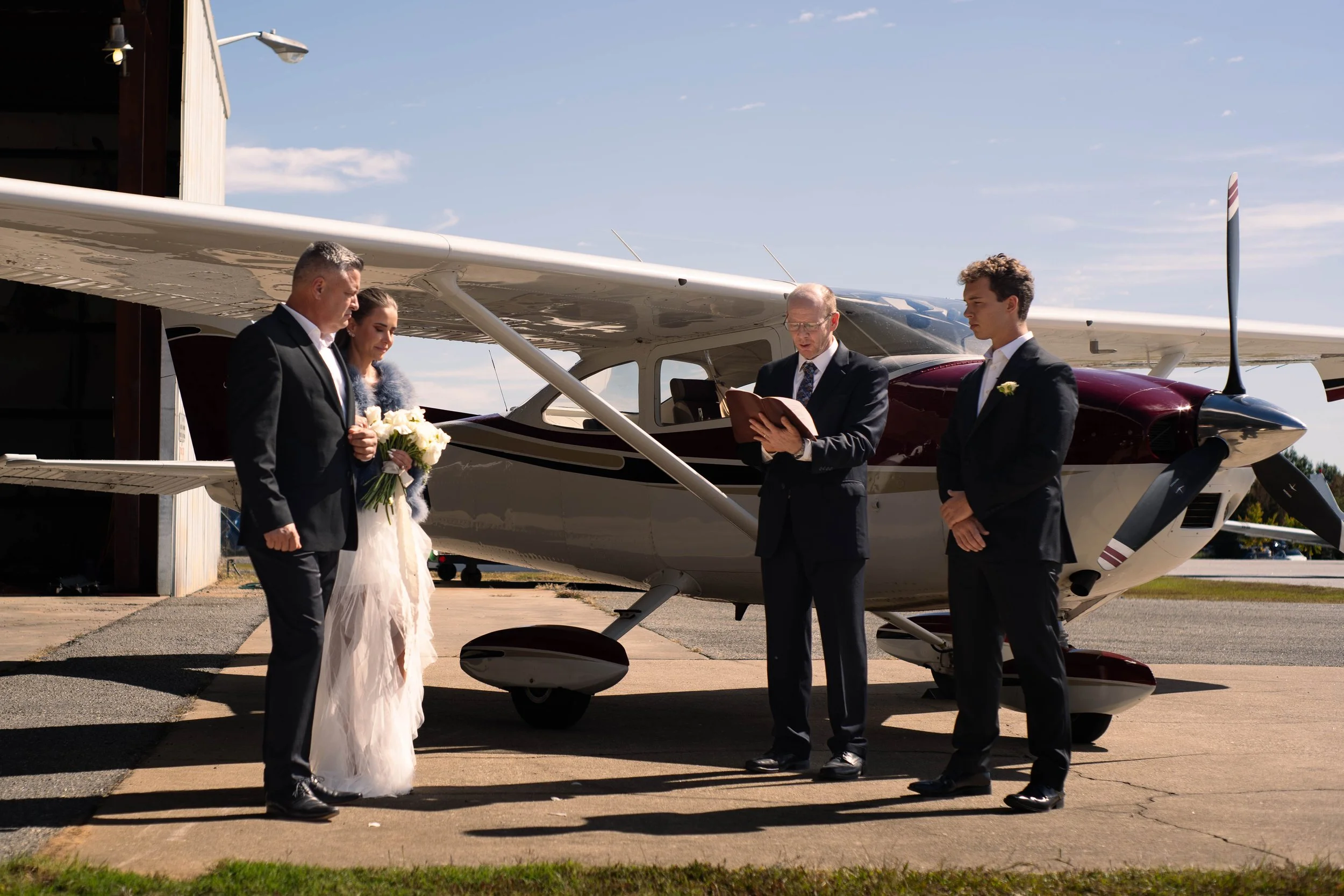 A wedding ceremony taking place on the tarmac beside a small airplane, with a officiant reading from a book while a bride and groom stand before him. The bride holds a bouquet of white flowers, and both are dressed in formal wedding attire. The groom