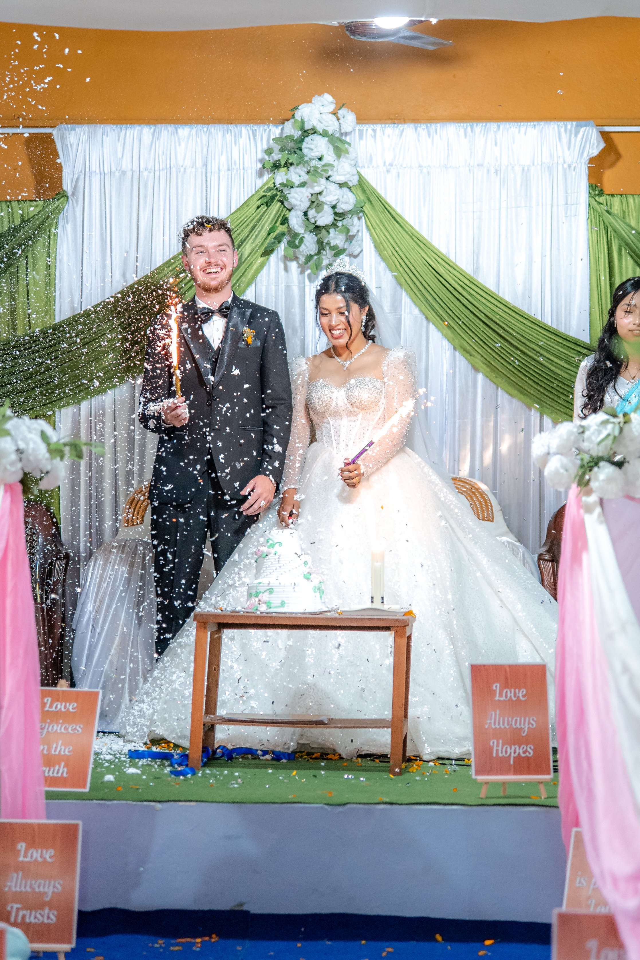 A newlywed couple celebrating their wedding with a sparkler in front of a decorated backdrop, standing behind a wedding cake and surrounded by flower arrangements and signs with love-related messages.