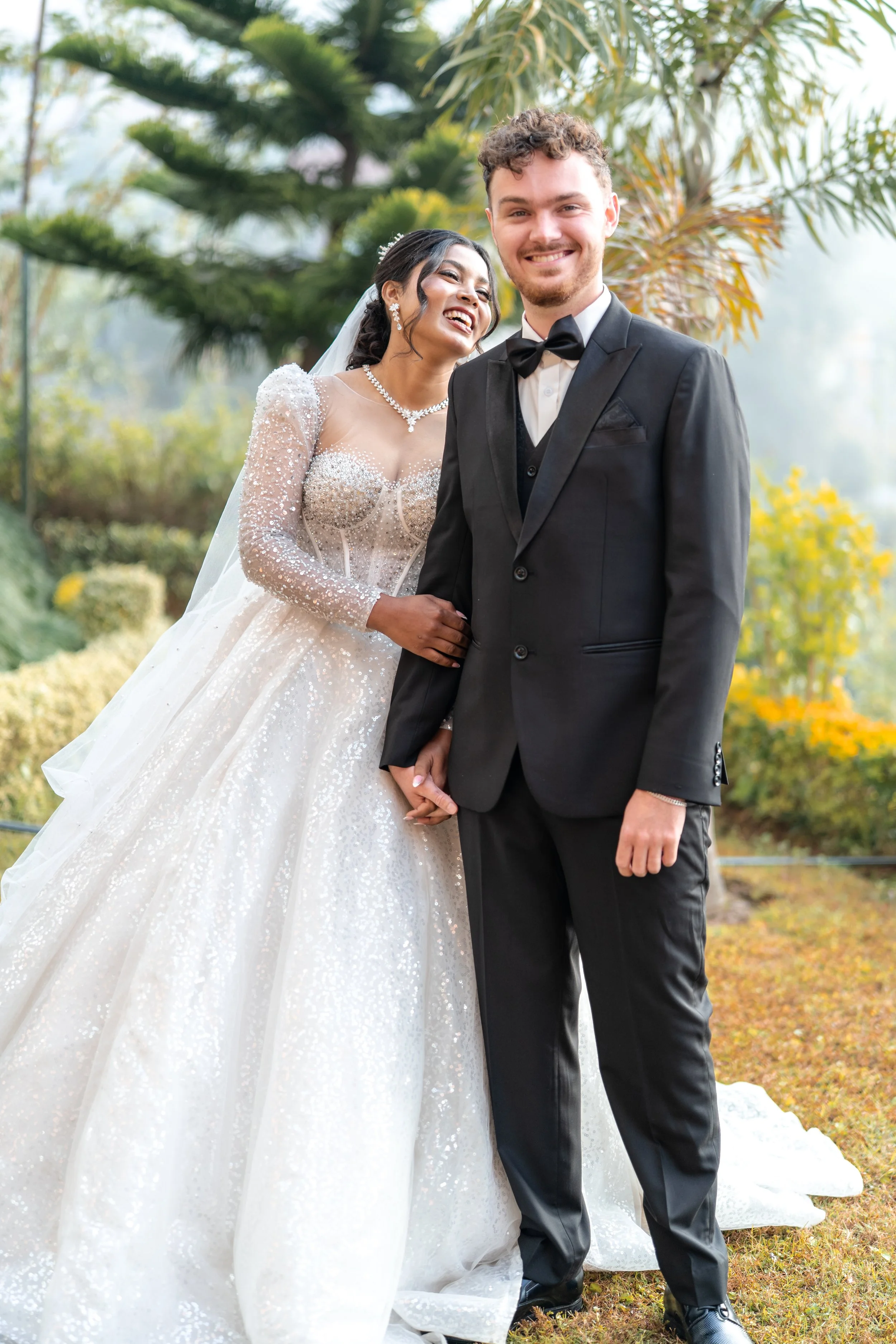 A bride and groom in wedding attire standing outdoors, holding hands, smiling, with trees and greenery in the background.