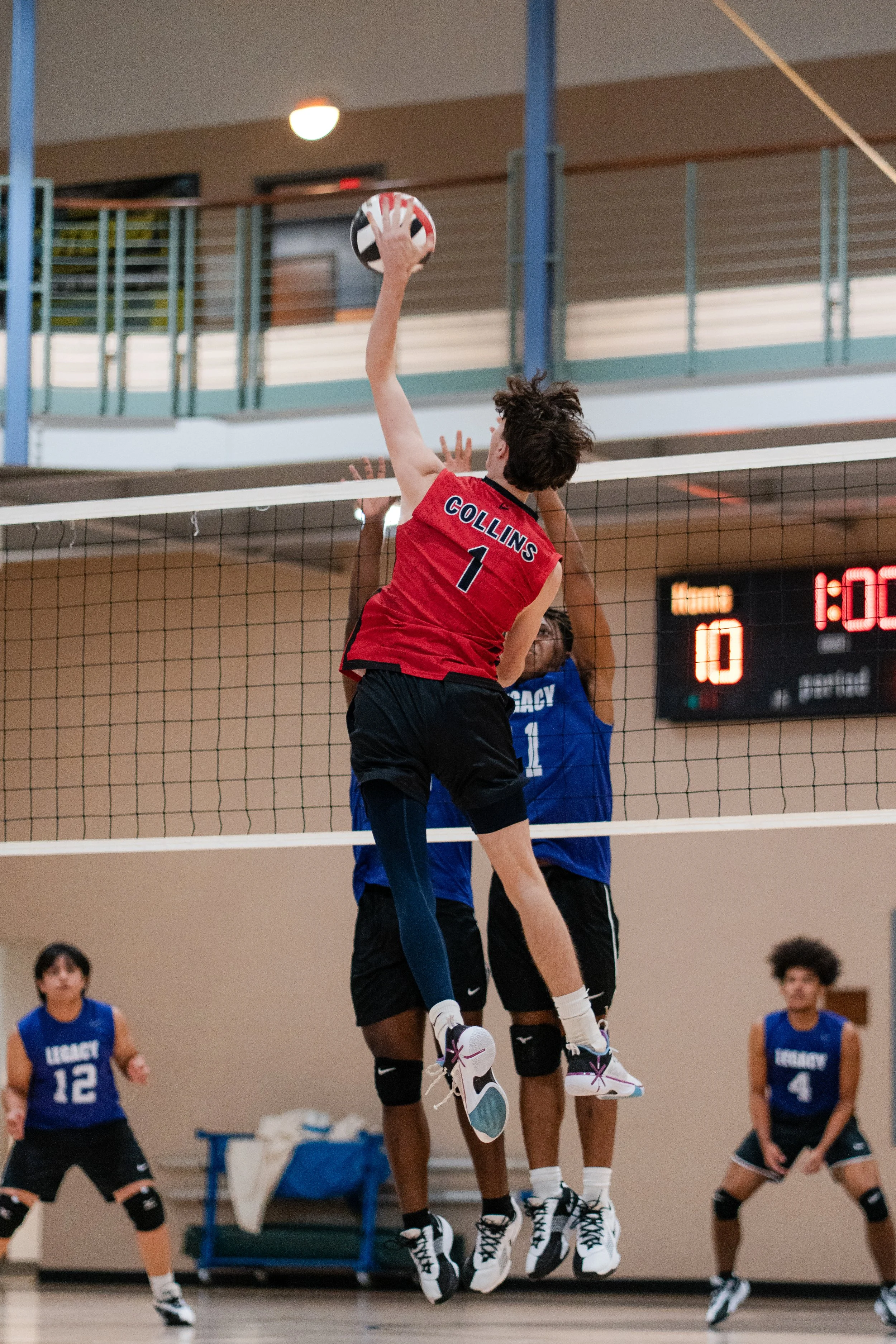 A volleyball player in a red jersey labeled 'Collins 1' is jumping to hit the ball at the net, while a player in a blue jersey labeled 'Legacy 1' attempts to block. Other players are visible in the background on the court.