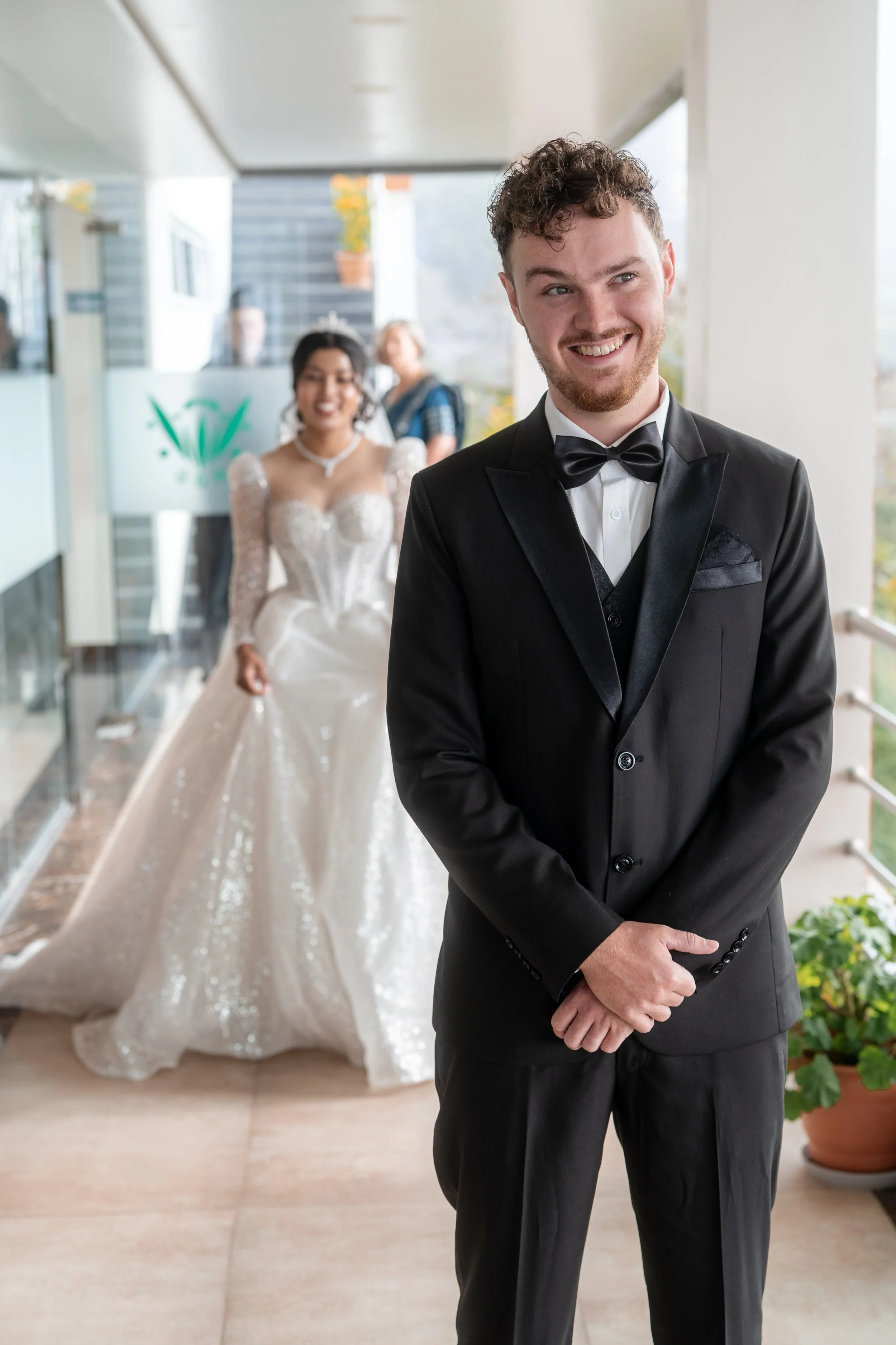 A groom in a black tuxedo with a bow tie stands smiling in front, with a bride in a white wedding gown in the background, walking toward him on a balcony or corridor.