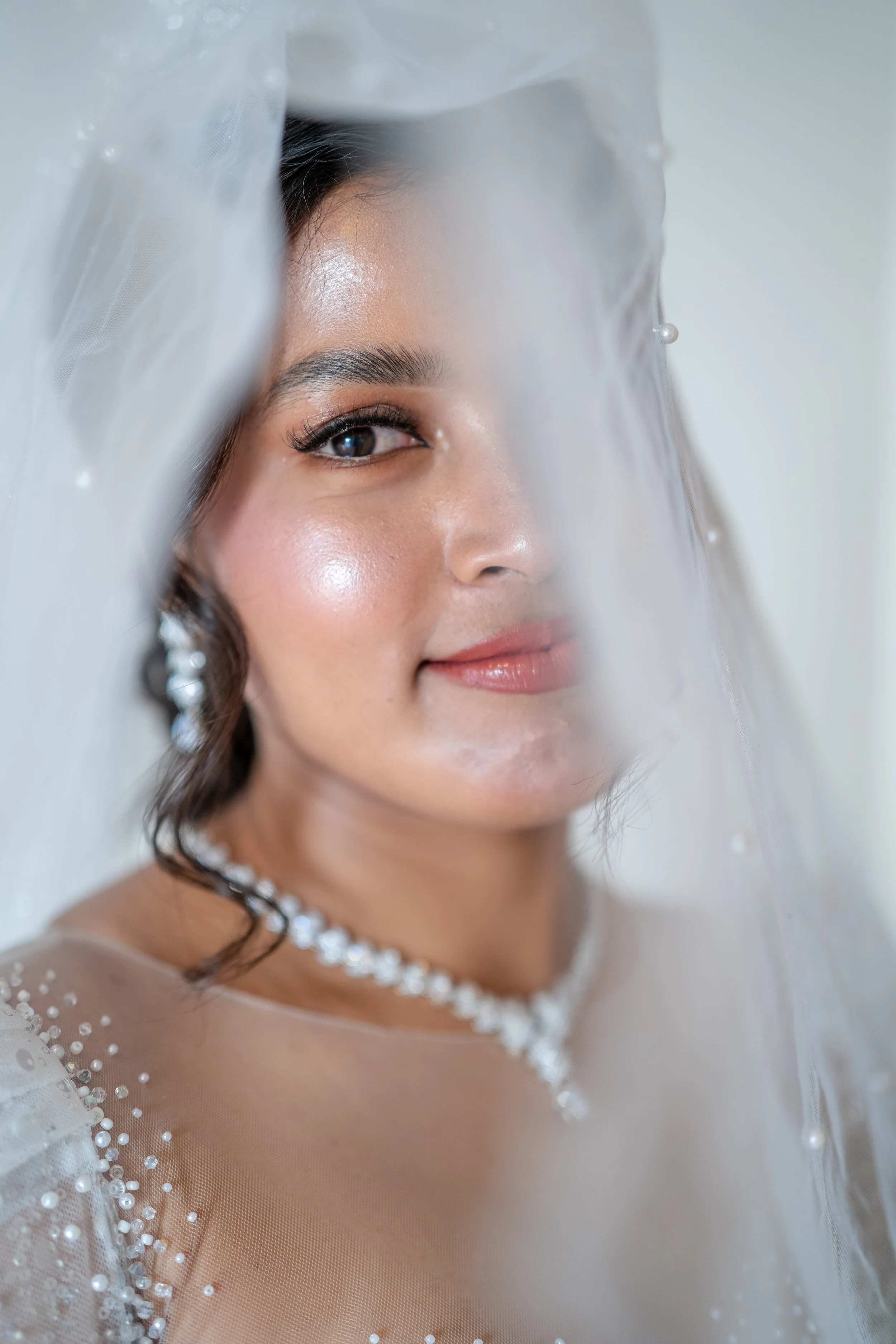 Close-up of a woman with wedding makeup and jewelry, partially obscured by a sheer veil.