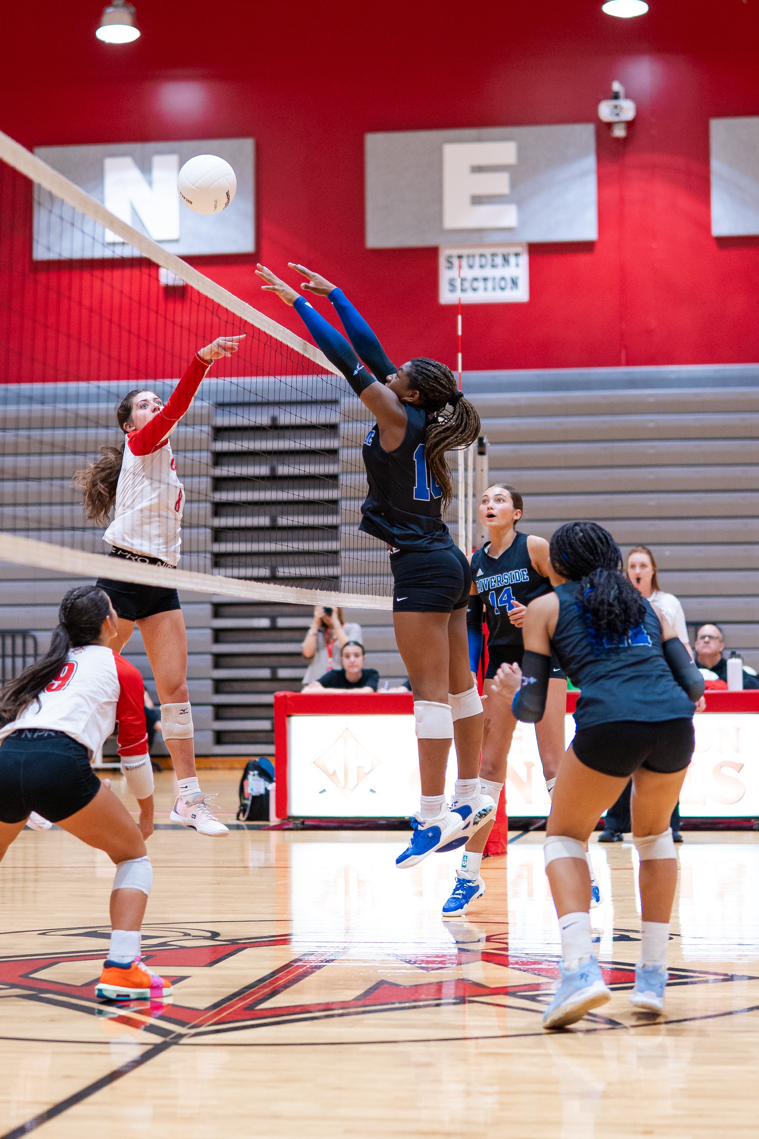 Female volleyball players in action during a match in a gymnasium, with one player jumping to spike the ball and others preparing to defend.