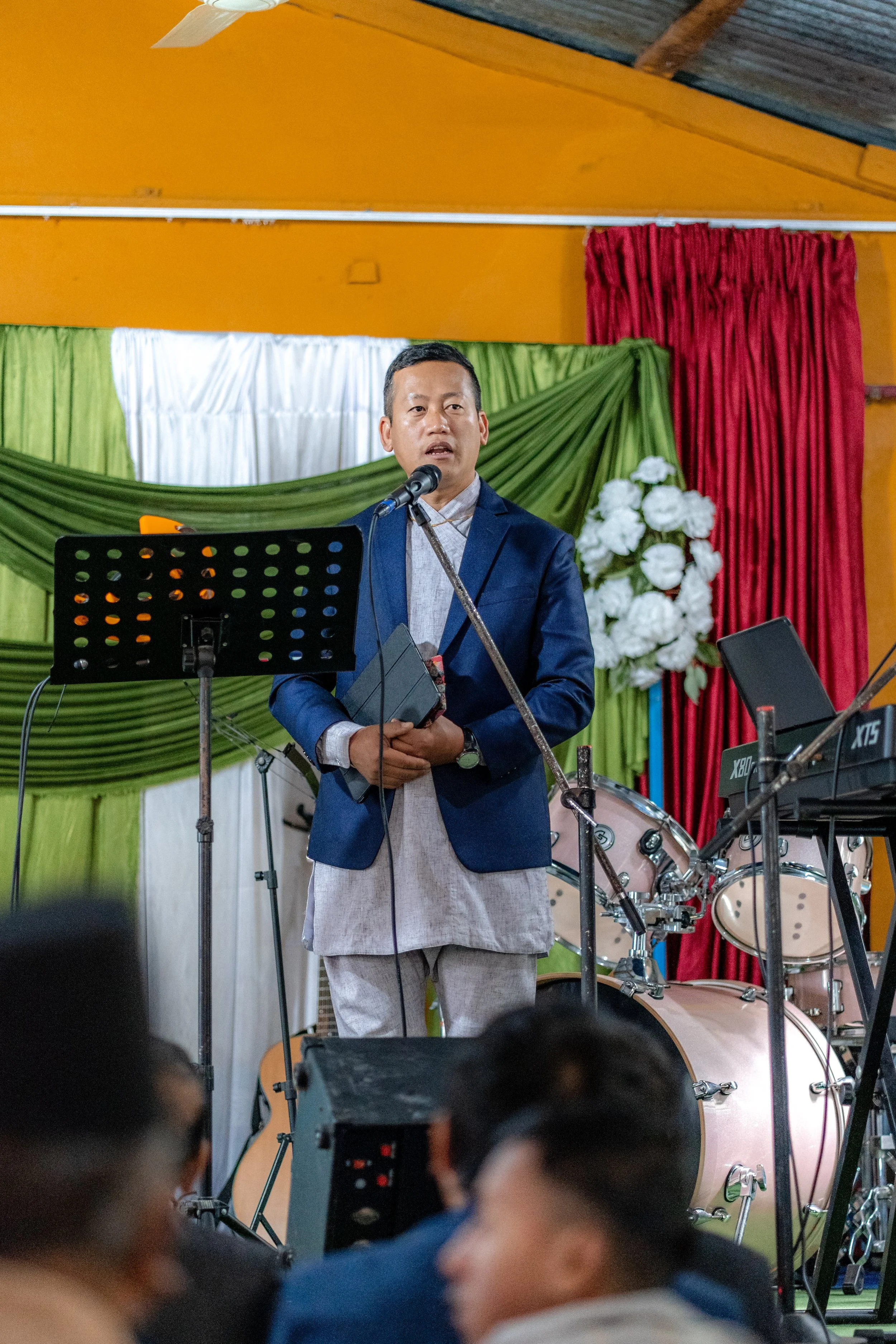A man in a blue blazer speaking into a microphone at an indoor event, standing in front of a colorful curtain backdrop with green, white, and red fabric and floral decorations.