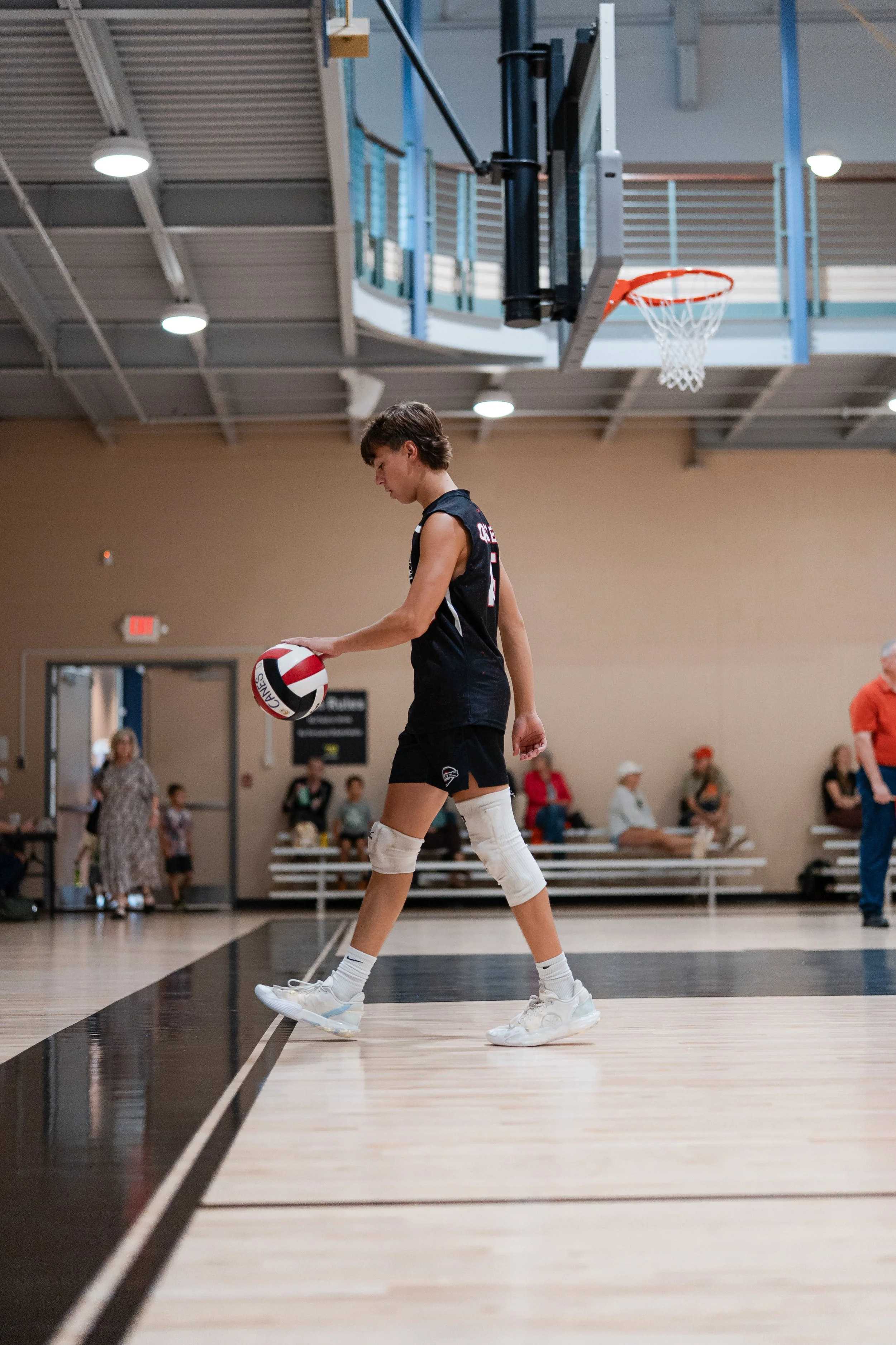 Young male volleyball player in black uniform with knee pads, holding a volleyball, walking on an indoor basketball court.