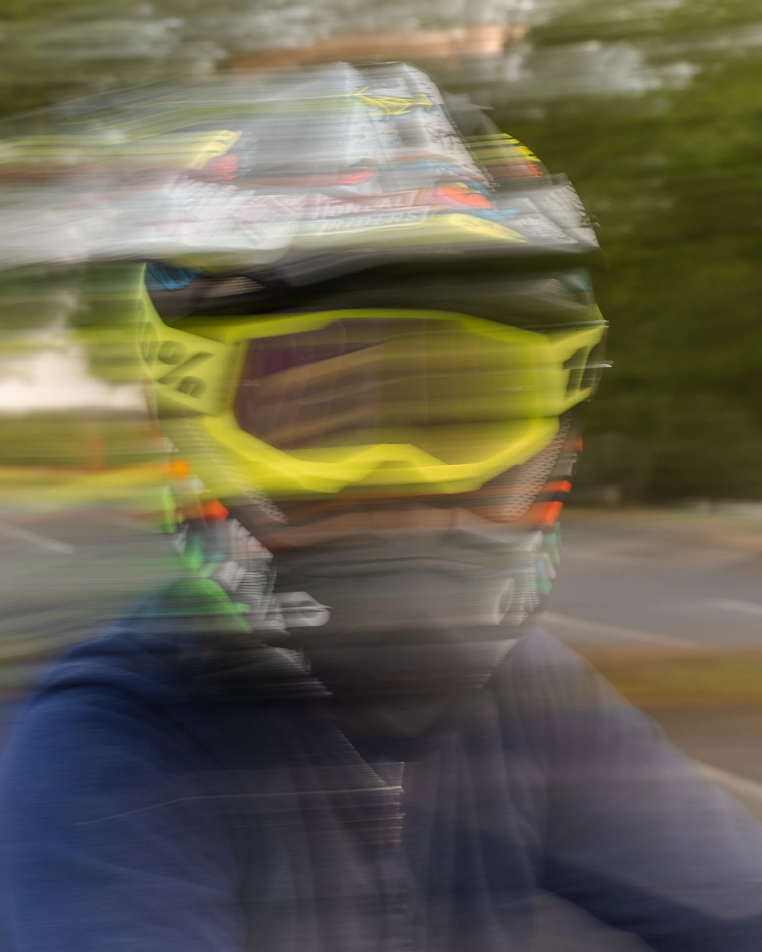 Blurry photo of a person wearing a neon yellow motorcycle helmet and a dark jacket, outdoors with trees in the background