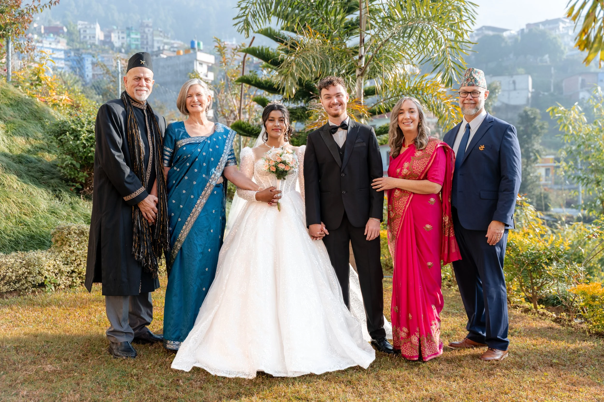 A wedding celebration outdoors with seven people of diverse ethnic backgrounds, including the bride in a white gown holding a bouquet, the groom in a black tuxedo, and five family members dressed in colorful traditional attire, standing in front of l