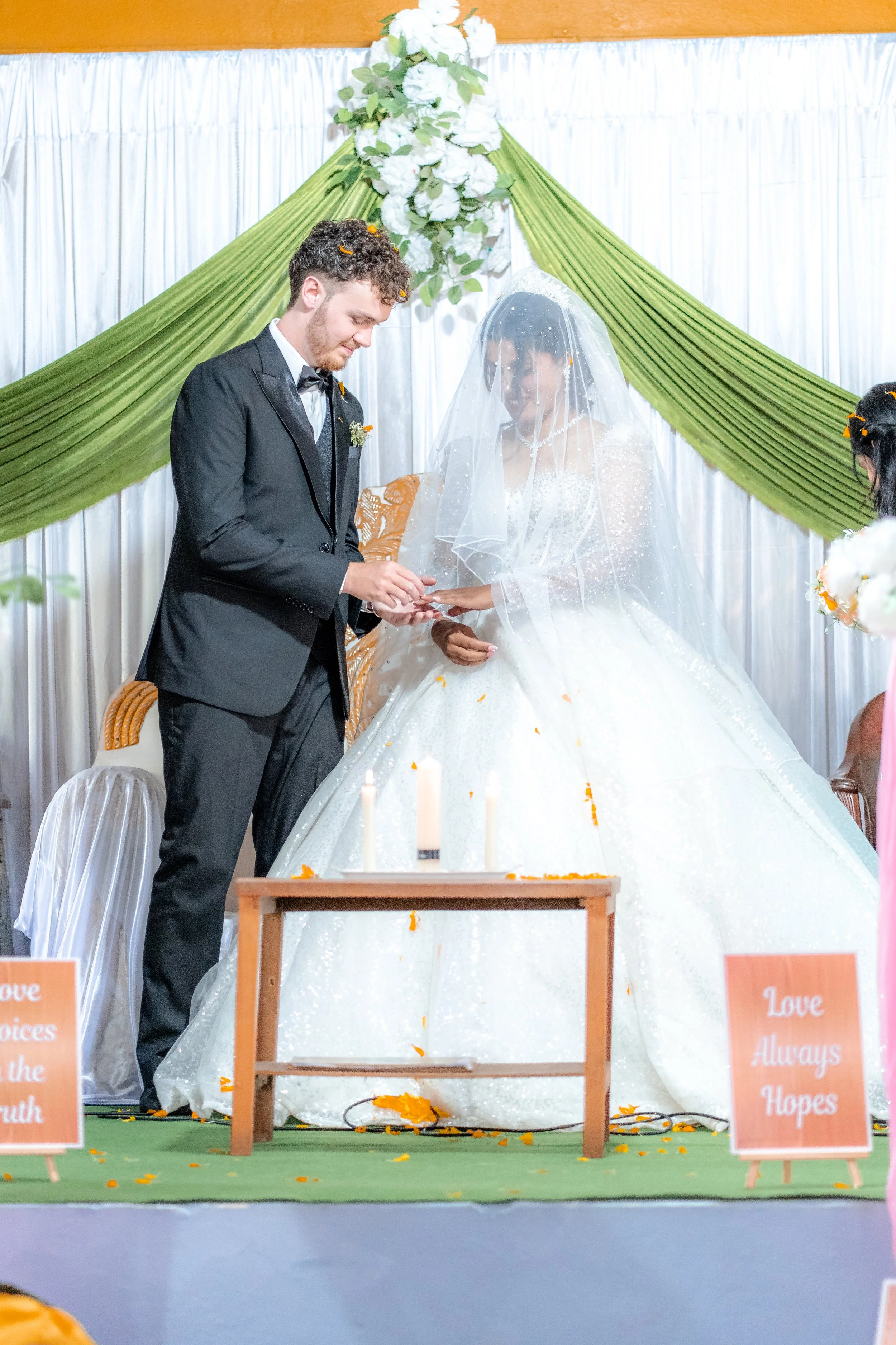 A bride and groom during their wedding ceremony, with the groom placing a ring on the bride's finger under a decorated arch with green drapes and white flowers. There are lit candles and flower petals on the floor, and signs that say 'Love Always Hop