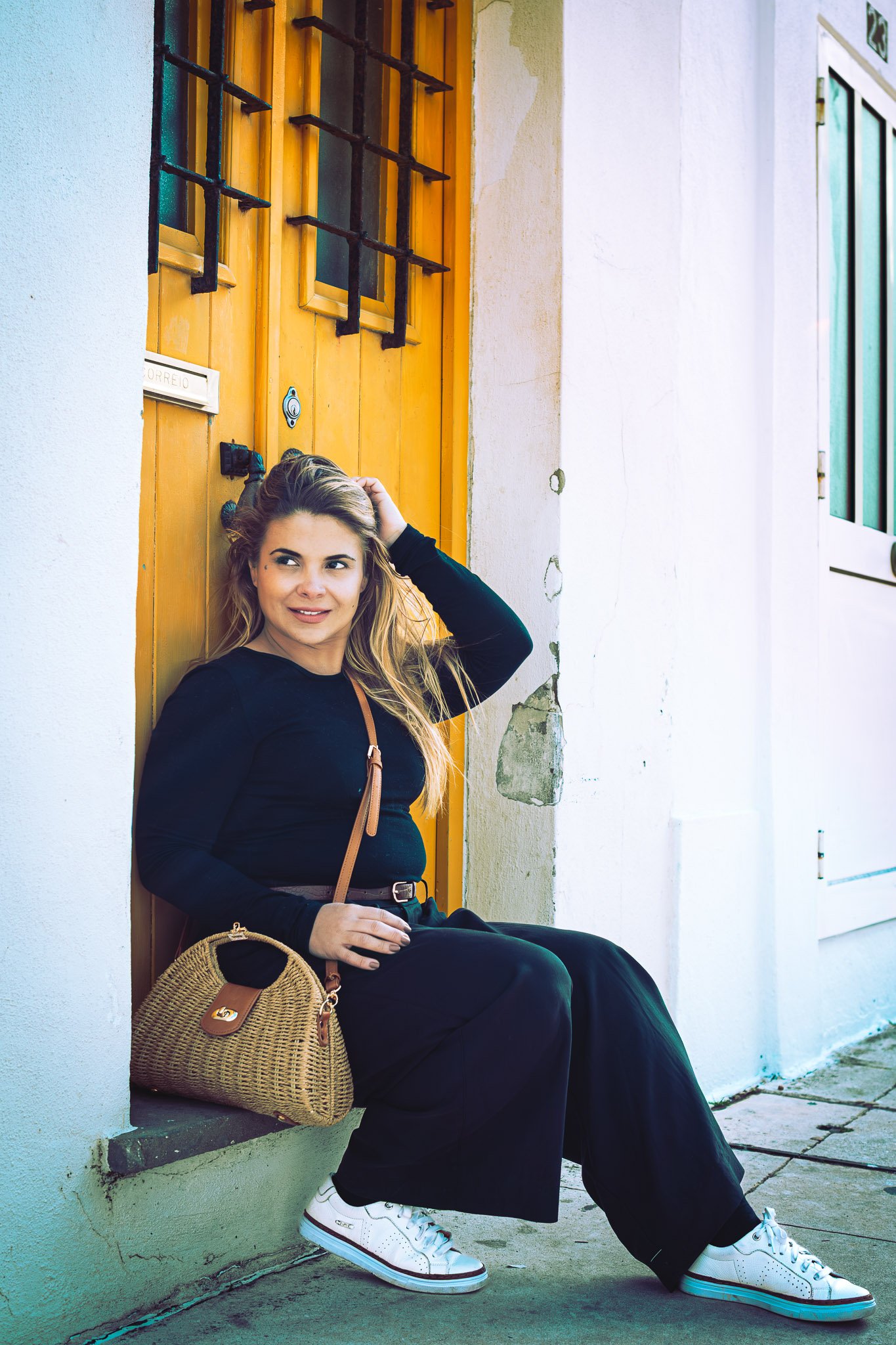 A young woman with long blonde hair, wearing a black long-sleeve shirt, black wide-leg pants, and white sneakers, sitting on a sidewalk against an orange door with security bars, holding her head with her right hand and looking to the side, while a woven handbag rests beside her.