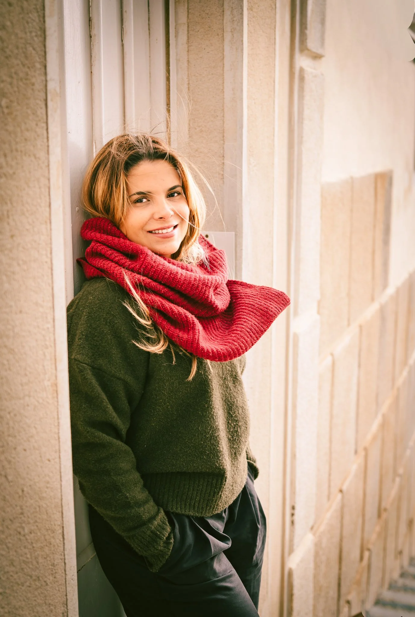 Young woman with blonde hair, wearing a green sweater and a red scarf, smiling and leaning against a beige wall outdoors.