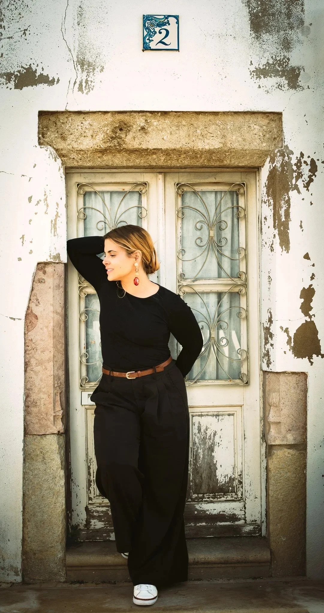 A woman with short hair, wearing a black top and black pants, stands in front of a weathered door with a small blue square sign displaying the number 2. She is smiling with her eyes closed, one hand behind her head, and the other hand resting on her hip. The background has peeling paint and exposed bricks.