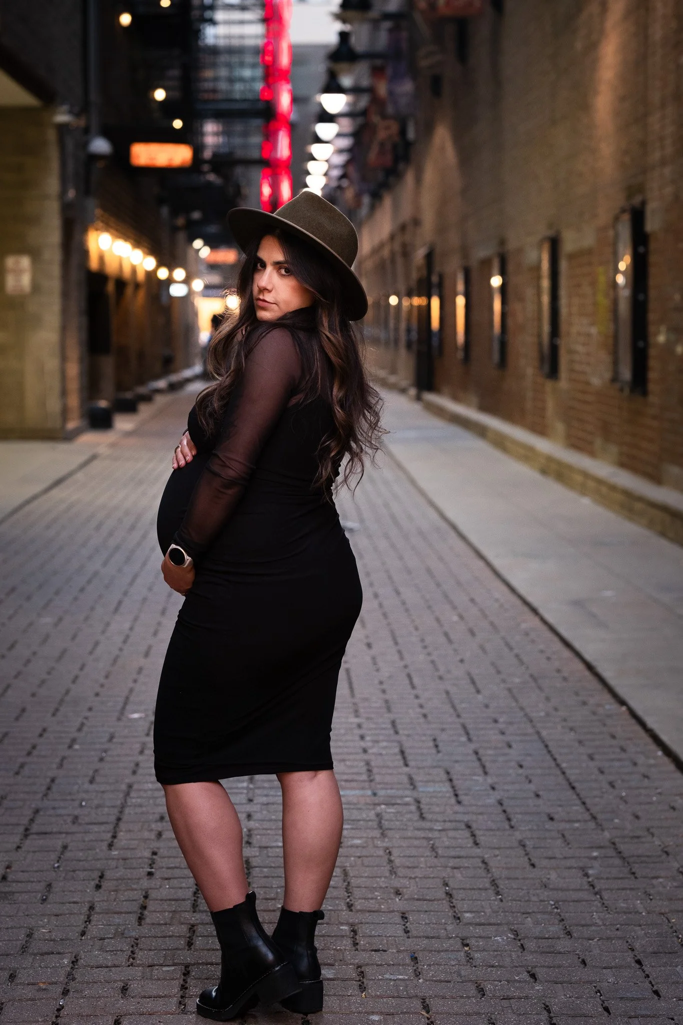 A pregnant woman with long wavy hair wearing a black dress, black boots, and a wide-brimmed hat stands on a city street at dusk, with brick buildings and illuminated streetlights in the background.
