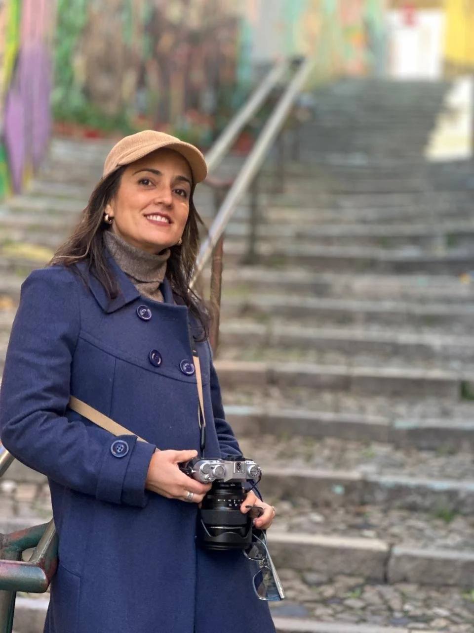 A woman in a blue coat and tan hat holding a camera, standing near colorful graffiti-covered stairs outdoors.