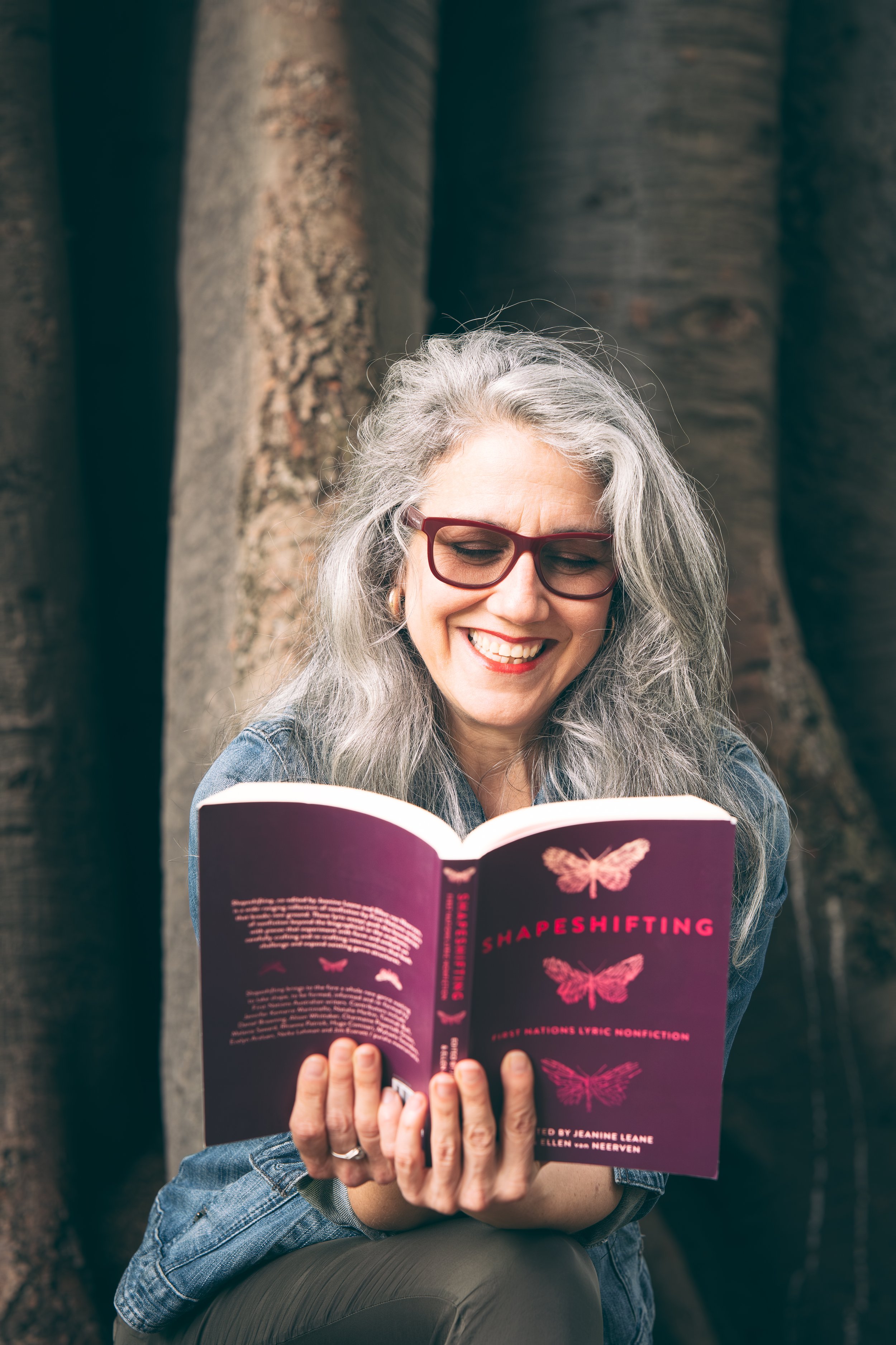 A smiling woman with gray hair and glasses reads a book titled 'Shapeshifting' outdoors near a large tree.