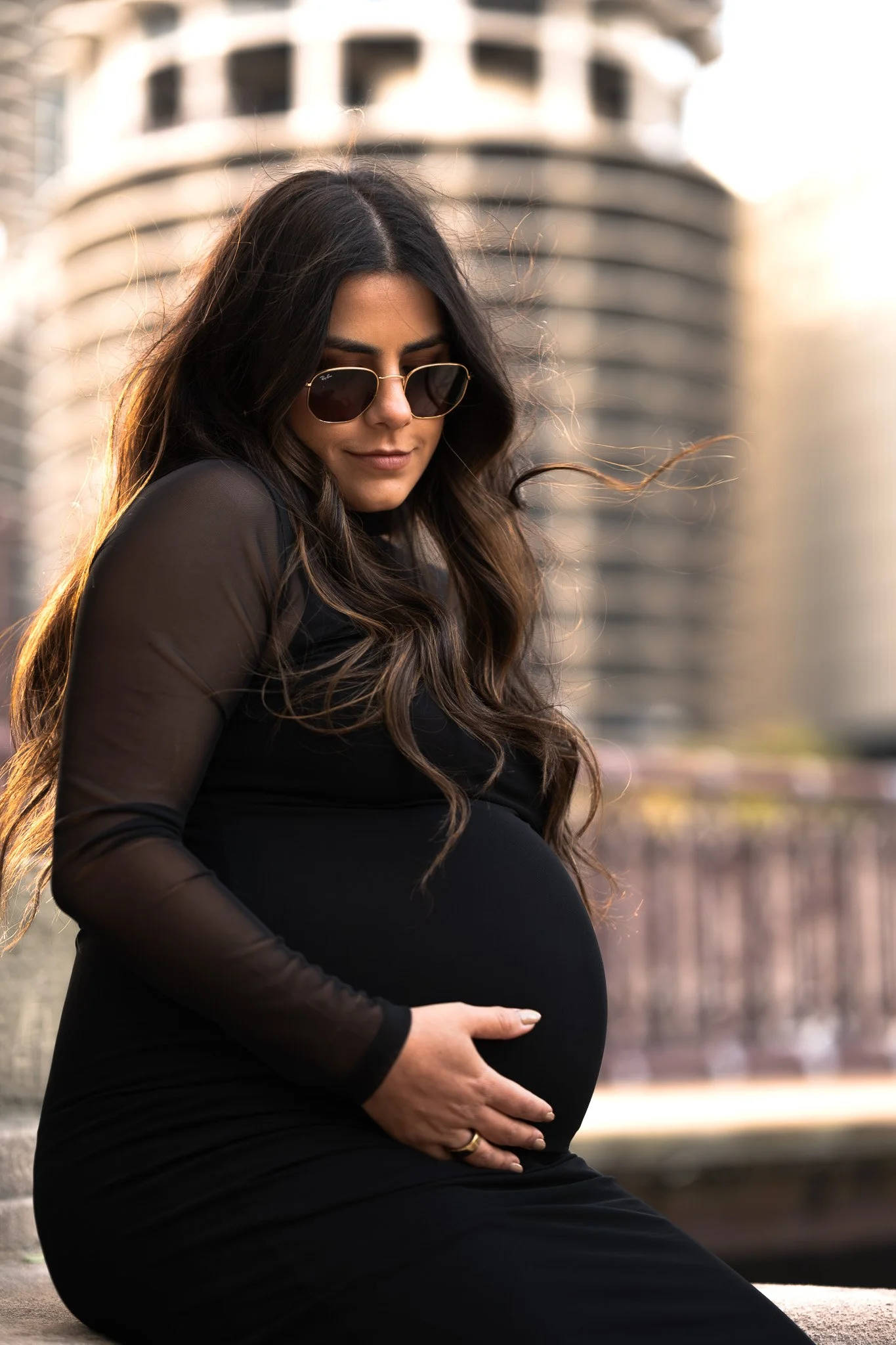 A pregnant woman with long dark hair wearing sunglasses and a black dress, holding her belly with one hand, sitting outdoors in front of a cityscape with tall buildings during sunset.