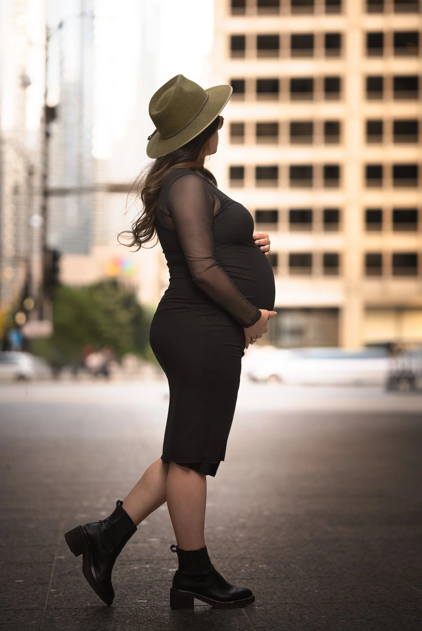 A pregnant woman with long curly hair, wearing a wide-brimmed hat, sunglasses, an  black sheer long-sleeve top, a black skirt, and black ankle boots, stands on a city sidewalk with her hands on her pregnant belly, looking to the side.
