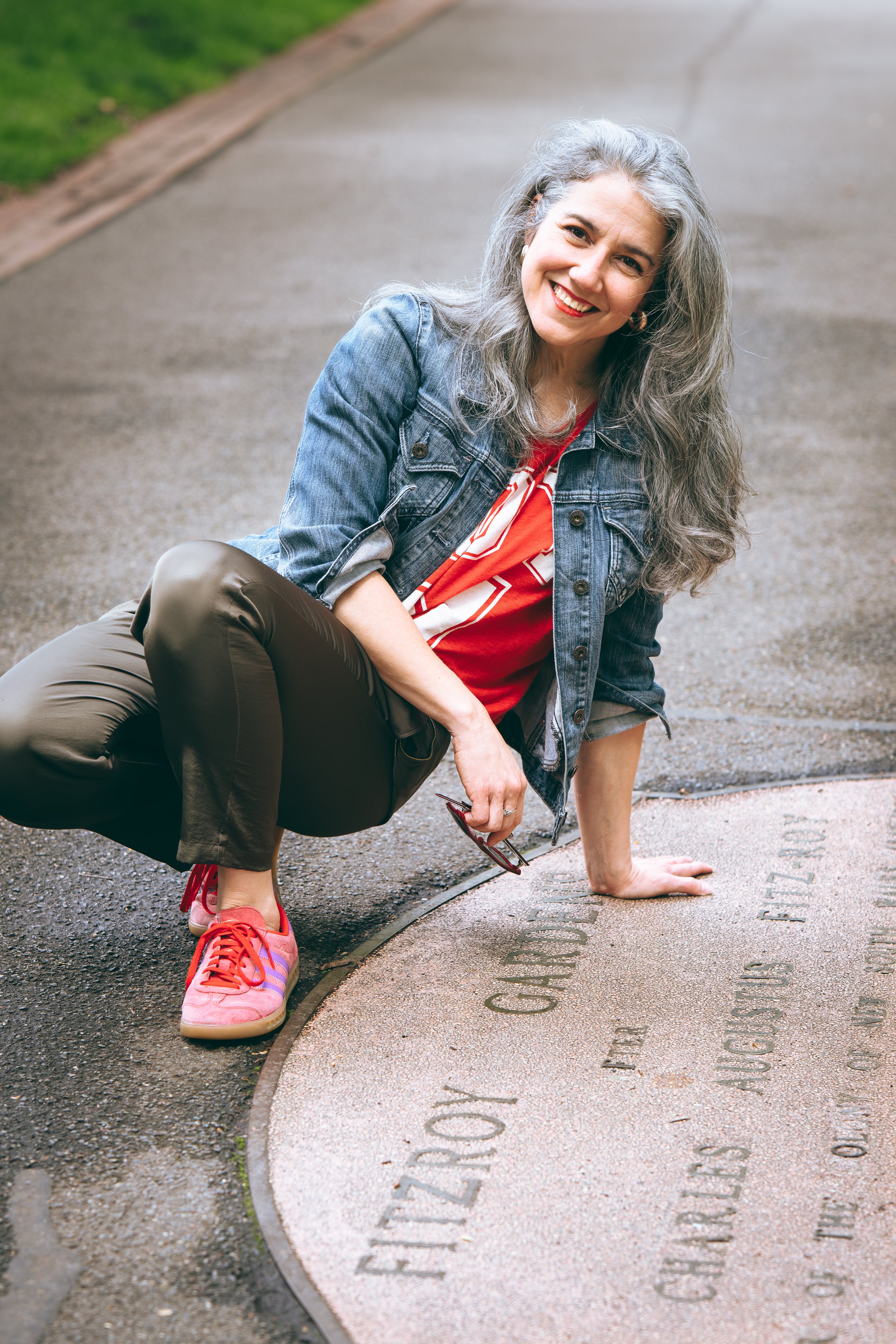 A woman with gray hair, smiling, crouching on a sidewalk next to a commemorative plaque, wearing a denim jacket, red shirt, gray pants, and pink sneakers.