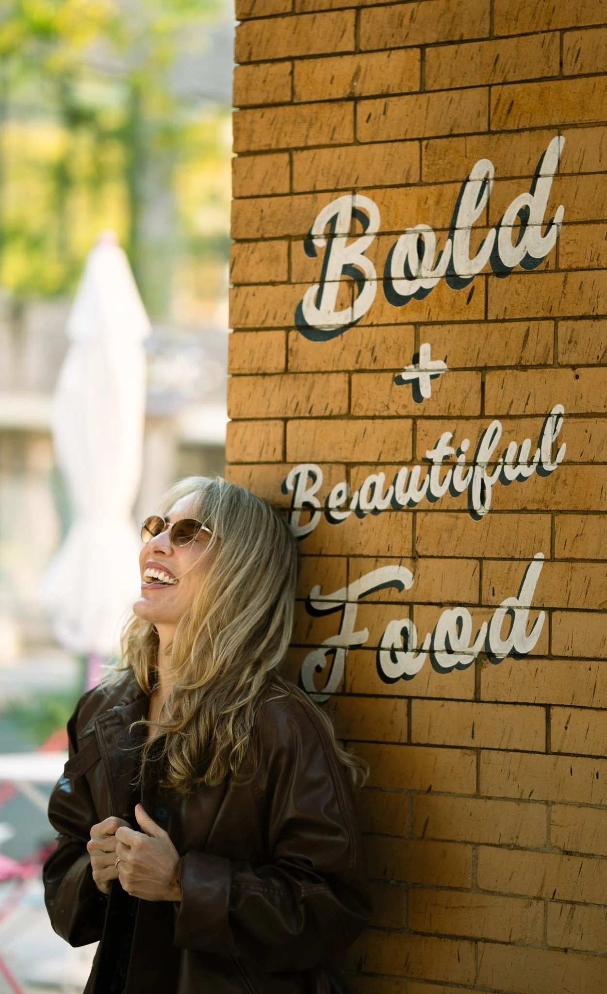 A woman with blonde hair, wearing sunglasses and a brown leather jacket, leaning against a brick wall with sunlight on her face, smiling and looking happy, with a mural on the wall that says 'Bold + Beautiful + Fearless'.