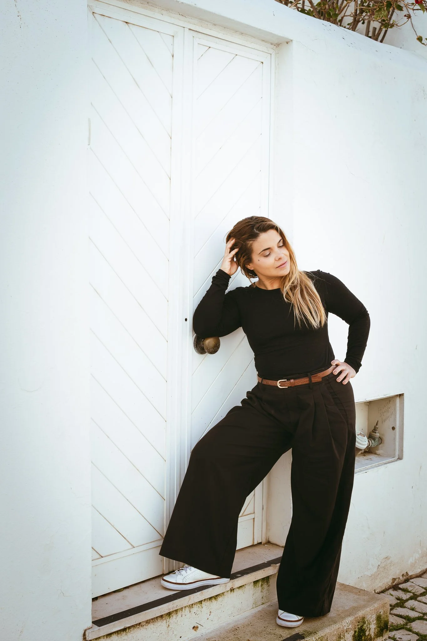 Woman in black long sleeve shirt and black wide-leg pants leaning against a white wall and door, with her eyes closed and hand in her hair.