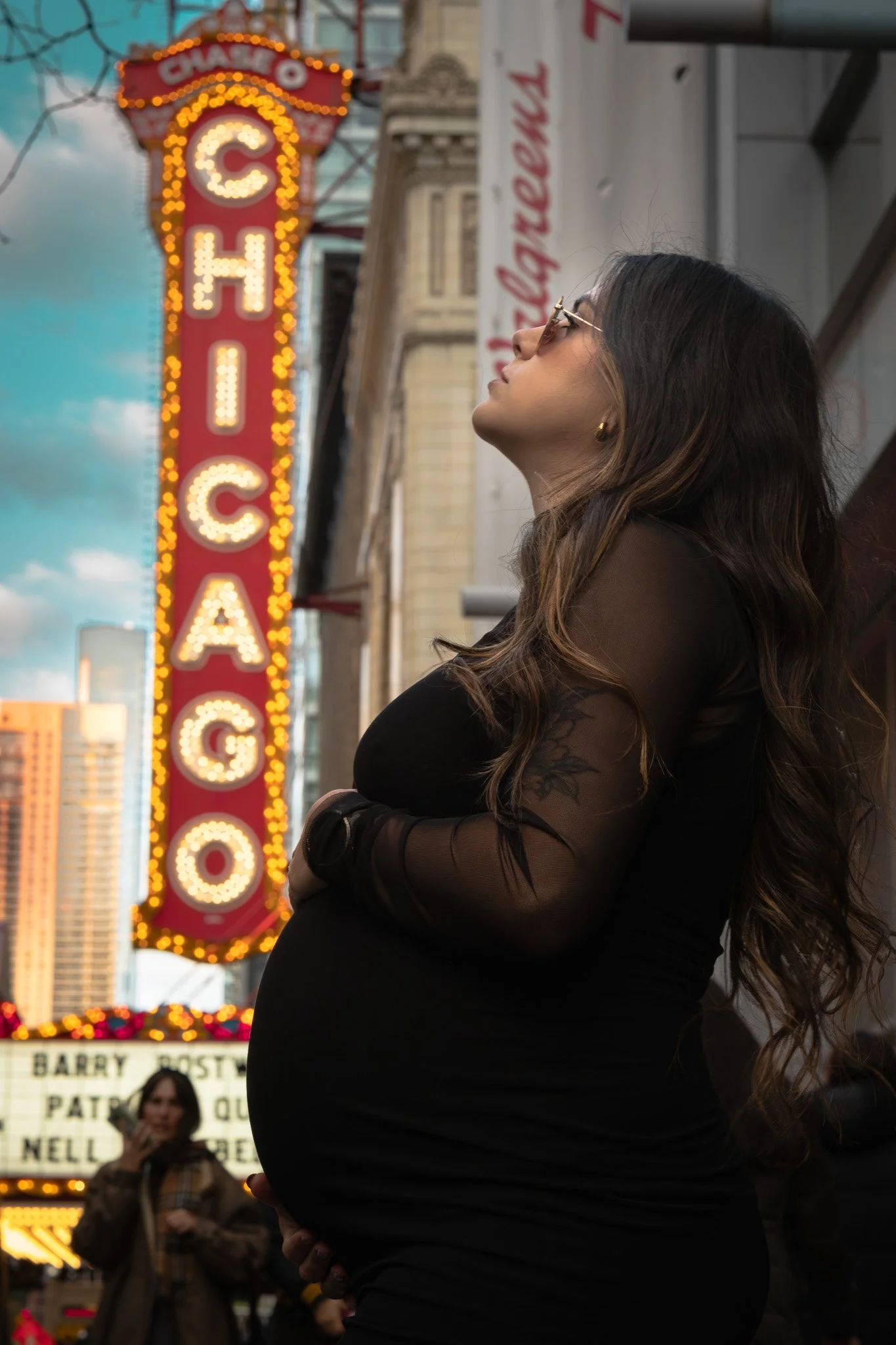 A pregnant woman with long wavy hair, wearing sunglasses and a black sheer top, stands outdoors on a city street with a large, illuminated 'CHICAGO' sign in the background, and a woman in a brown coat and scarf holding a phone in the distance.