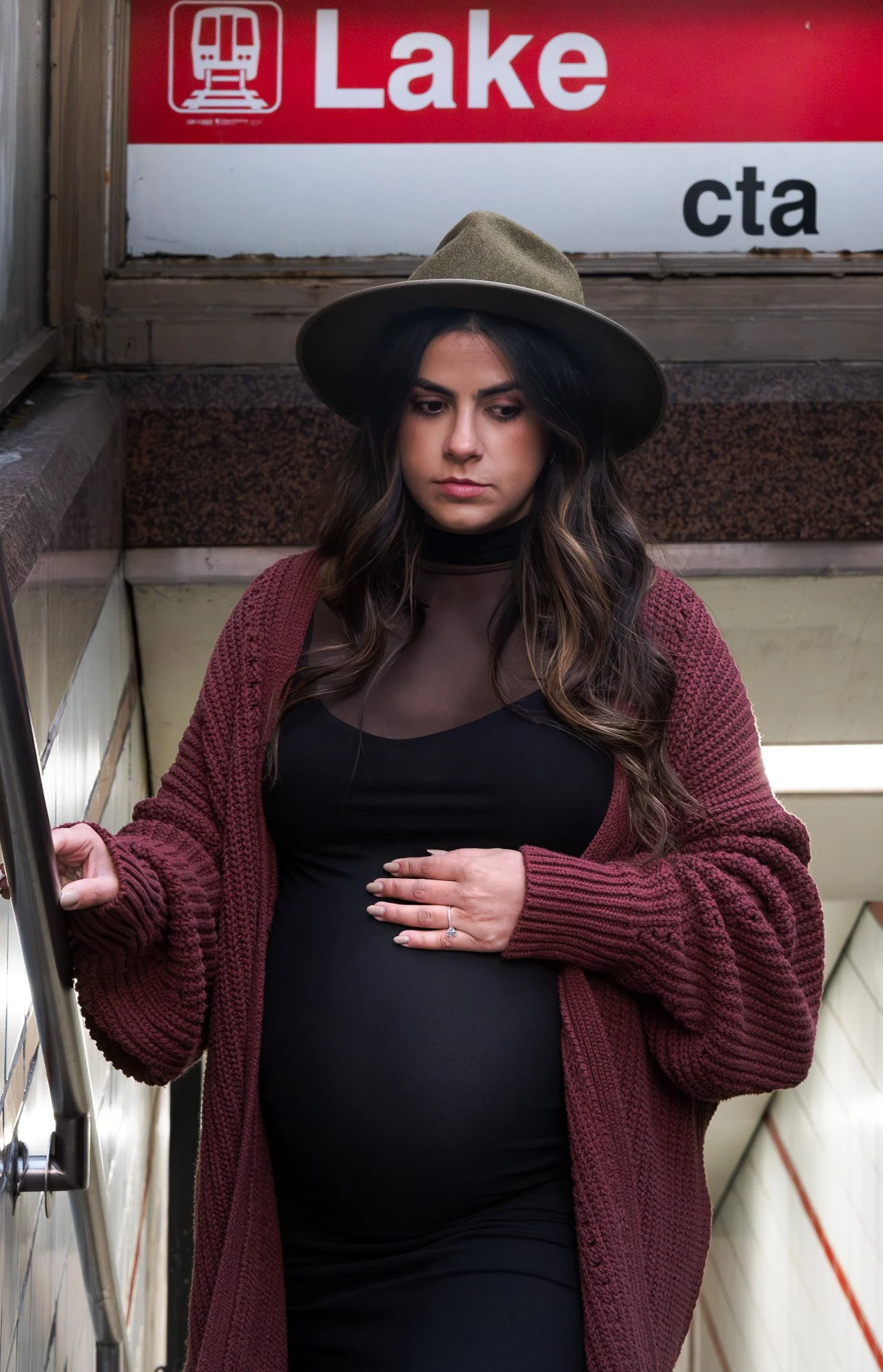 A pregnant woman with long dark hair wearing a wide-brimmed gray hat, black outfit, and maroon knit cardigan, standing on an escalator with a red and white