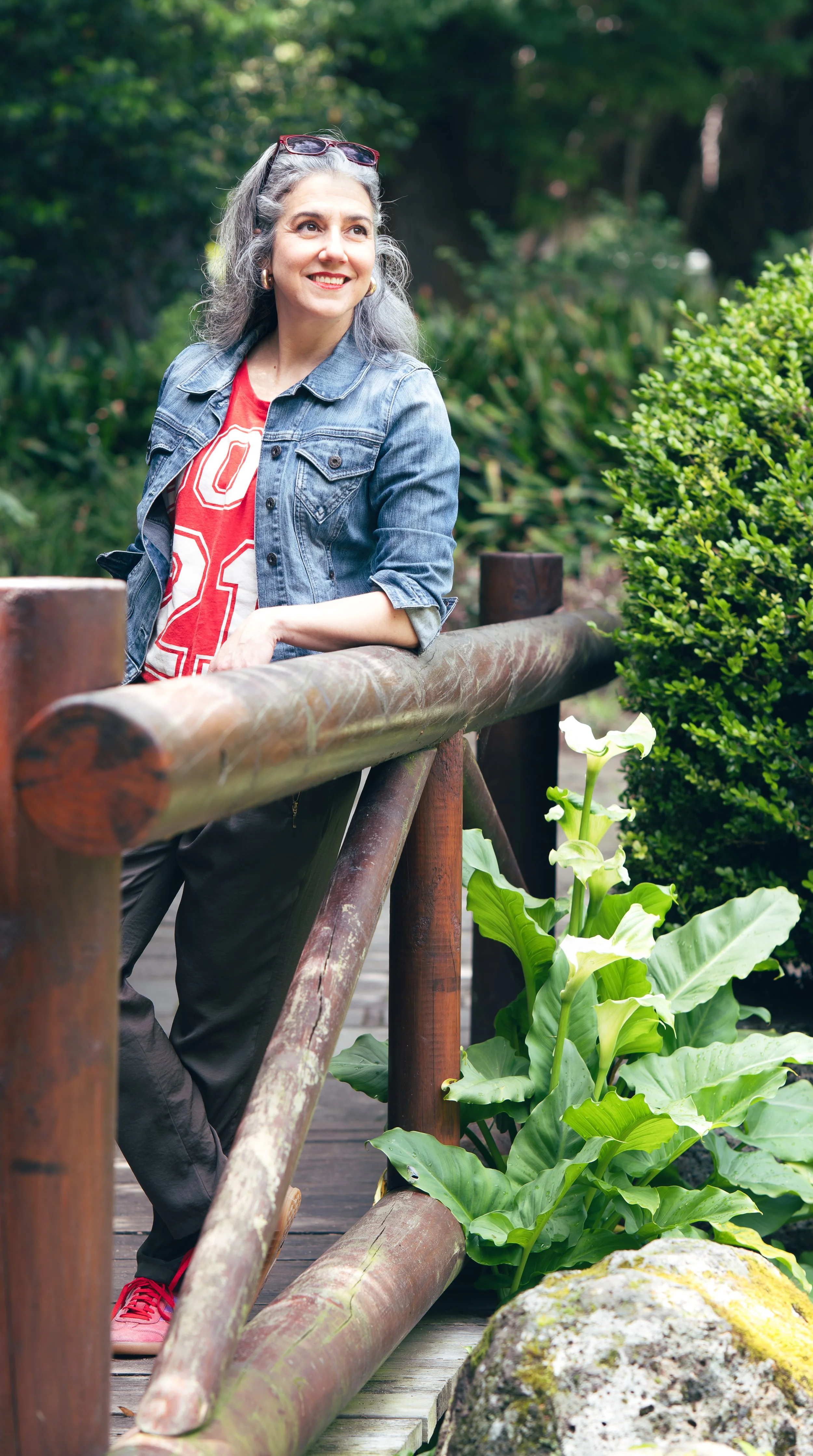 A woman with gray hair, wearing a denim jacket, red shirt, black pants, and red shoes, stands outdoors on a wooden bridge, smiling and looking to her left, with green foliage and white flowers in the background.