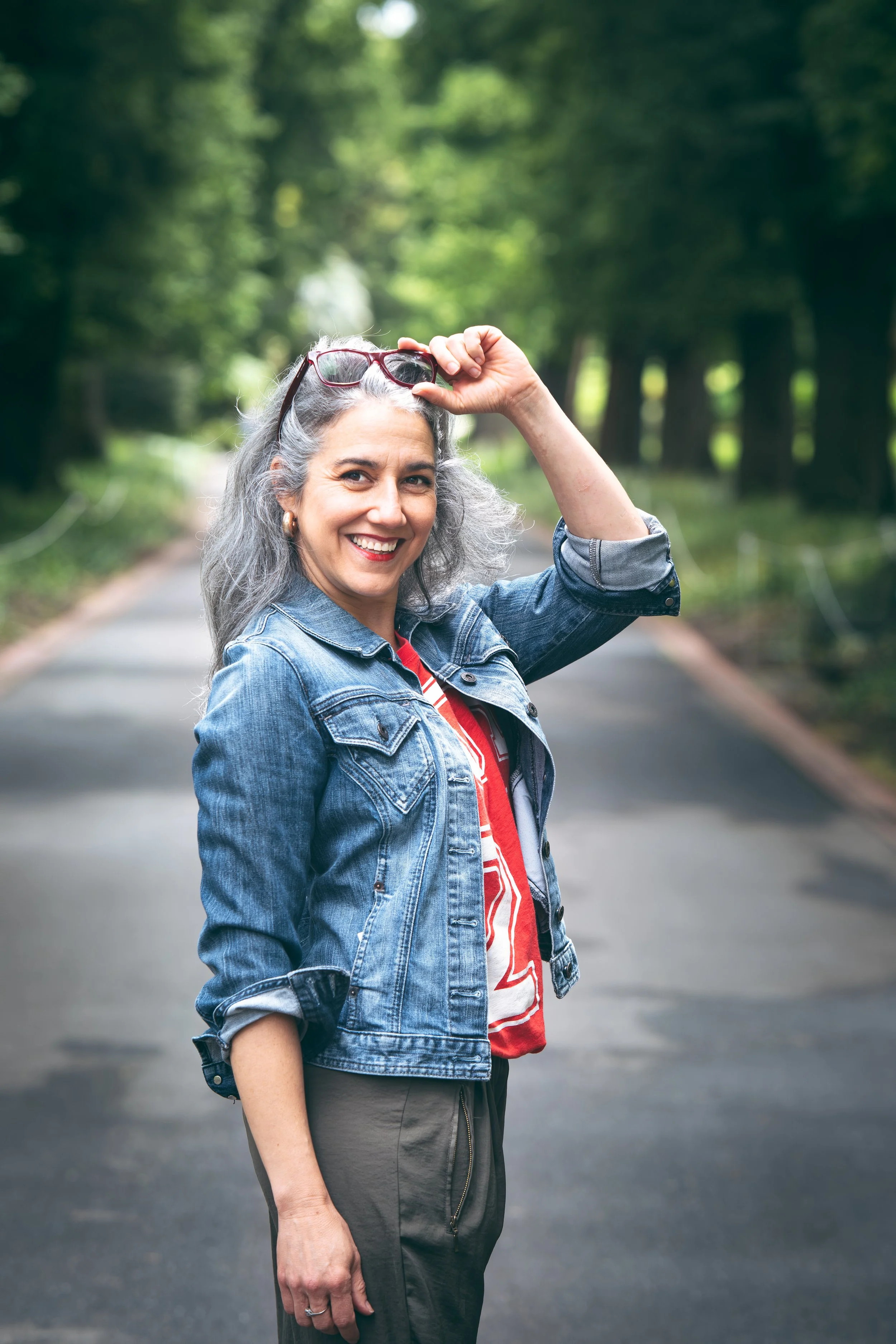 A woman smiling and adjusting her sunglasses while standing on a paved road in a green, wooded outdoor setting.
