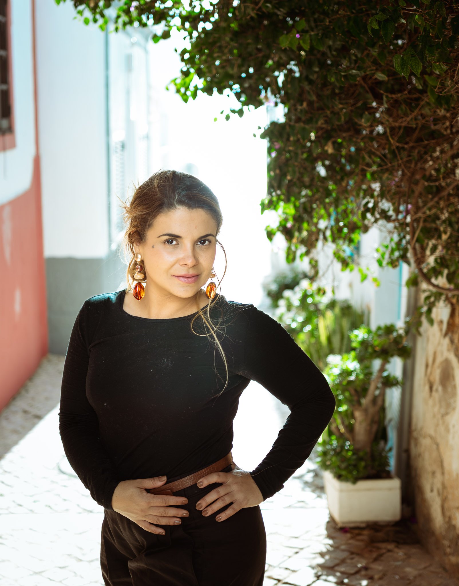 A woman standing outdoors in a narrow cobblestone alleyway with green foliage overhead. She has light brown hair and is wearing a black long sleeve top and earrings, with one hand on her hip looking at the camera.